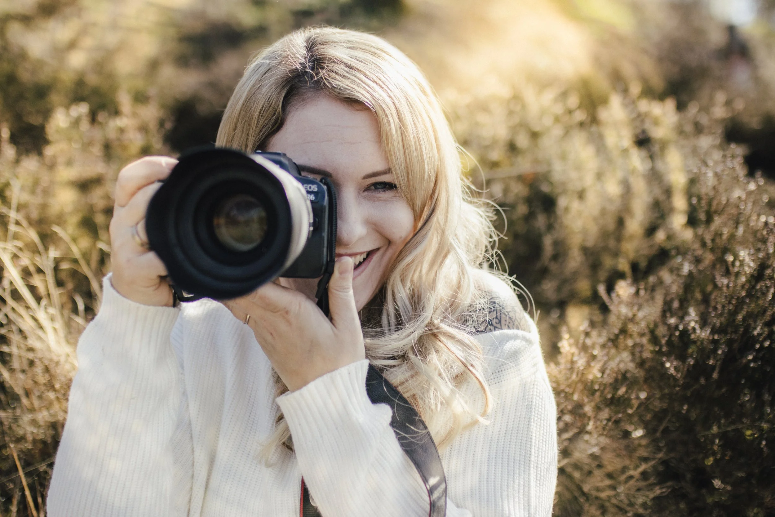Junge Frau mit blondem Haar, die eine Kamera auf ein Outdoor-Landschaft richtet, bei Sonnenlicht in herbstlicher Umgebung fotografierend.