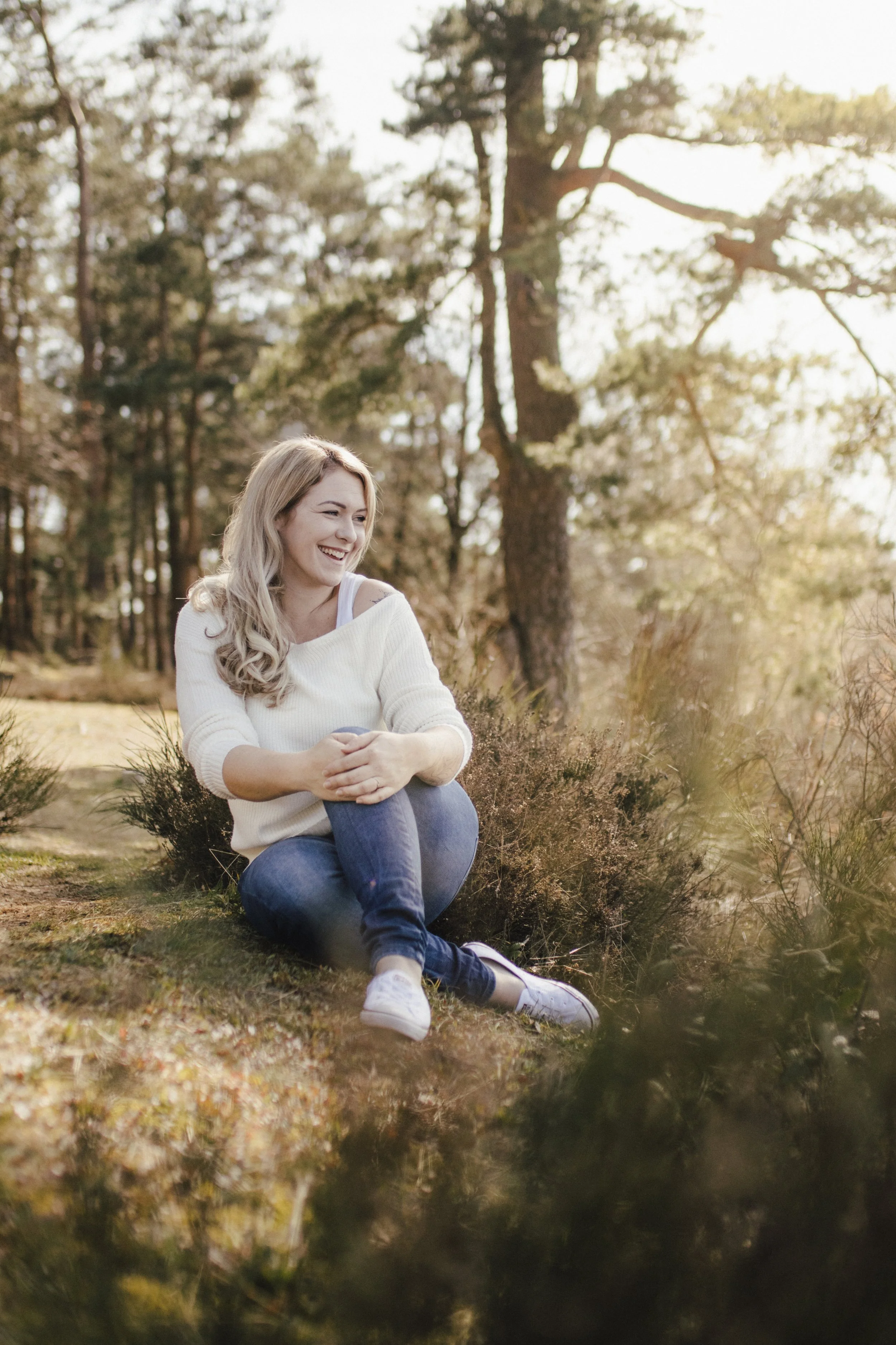Lächelnde junge Frau sitzt im Wald, umgeben von Bäumen und Herbstlaub, bei Sonnenuntergang.