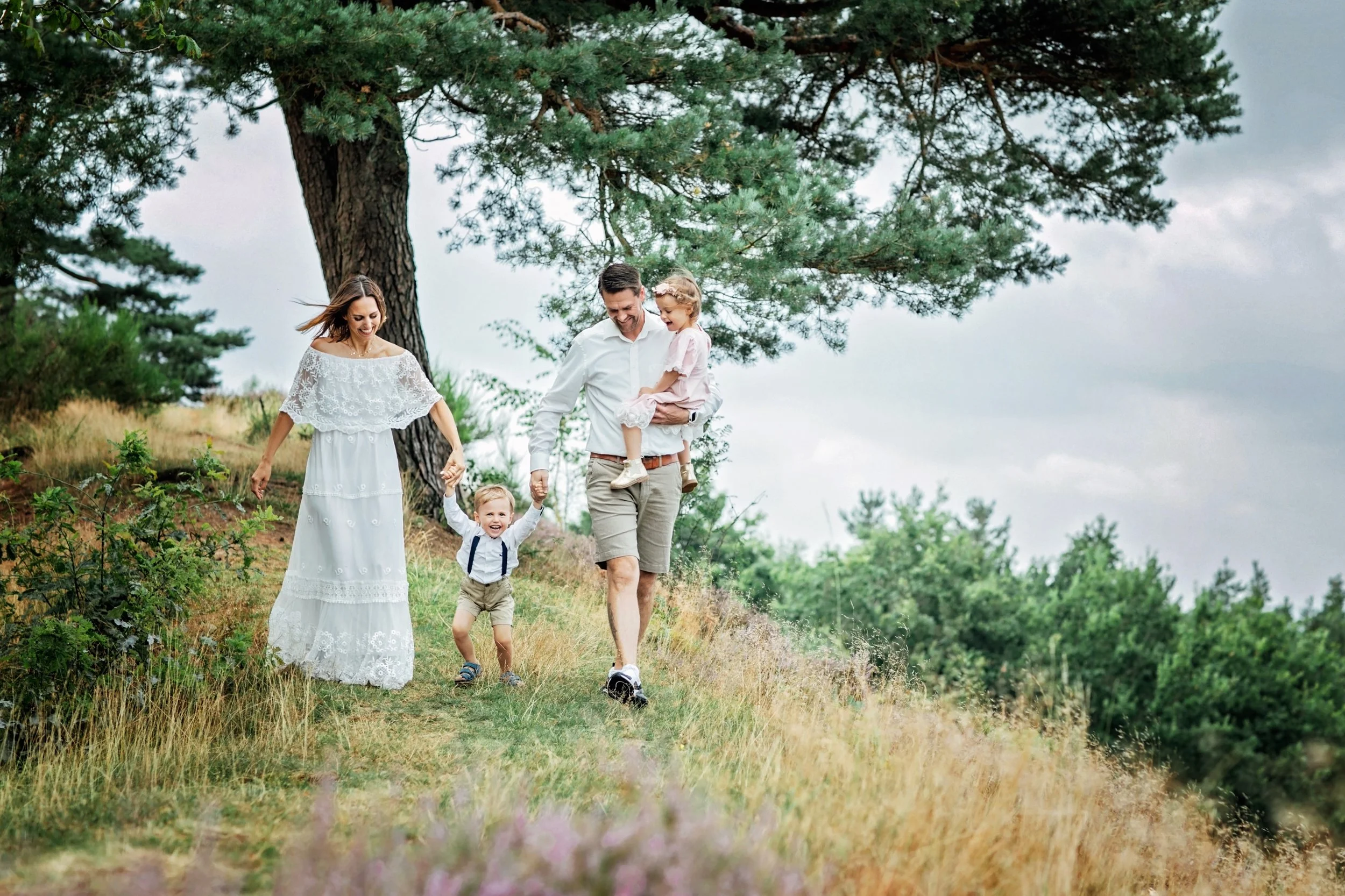 Eine Familie mit zwei Erwachsenen und zwei Kindern wandert auf einem Weg in der Natur, umgeben von Bäumen und Gras, bei bewölktem Himmel.