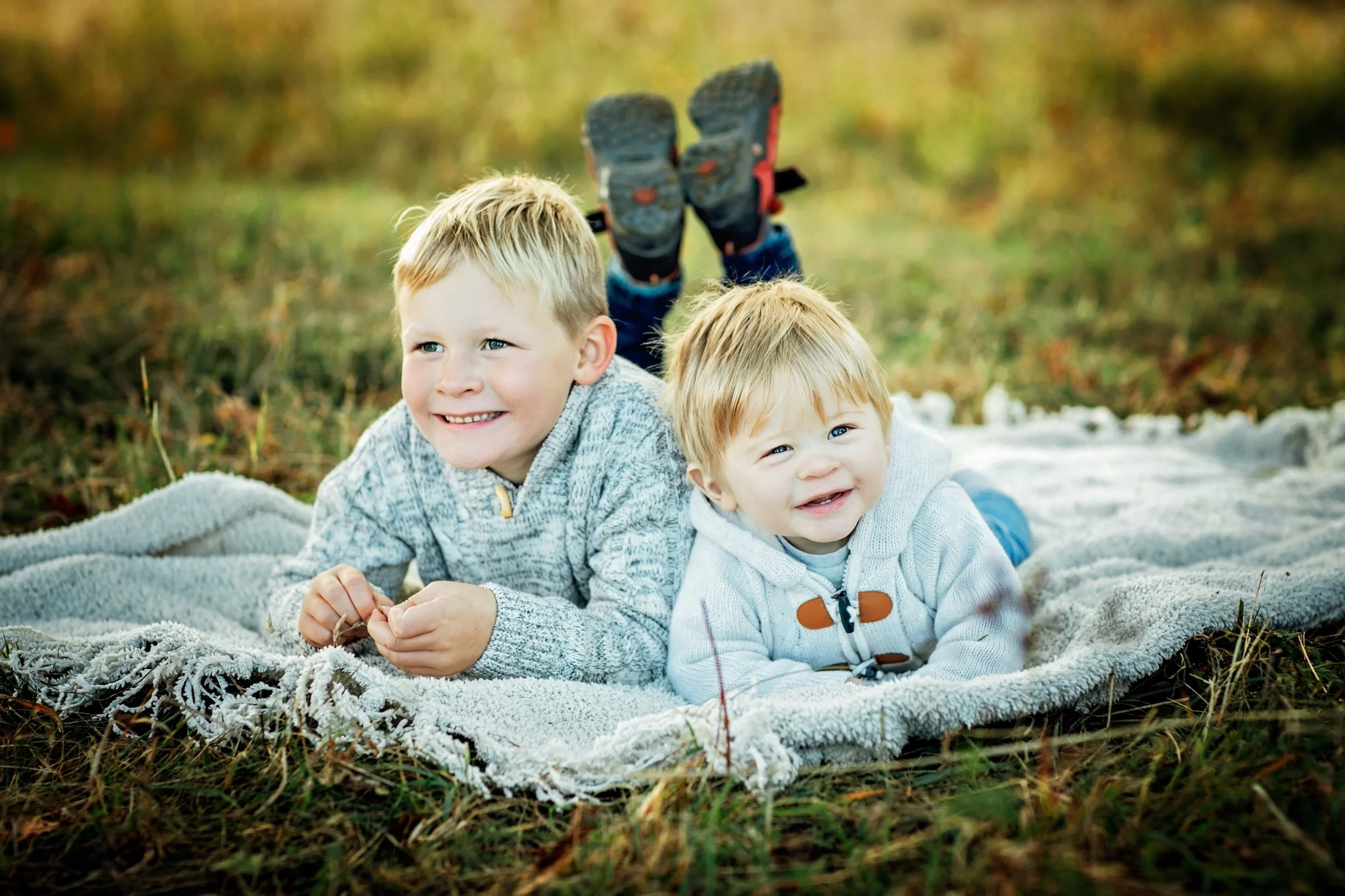 Zwei junge Jungen liegen auf einem Teppich im Freien, umgeben von Gras und Herbstlaub, lächeln und schauen in die Kamera.