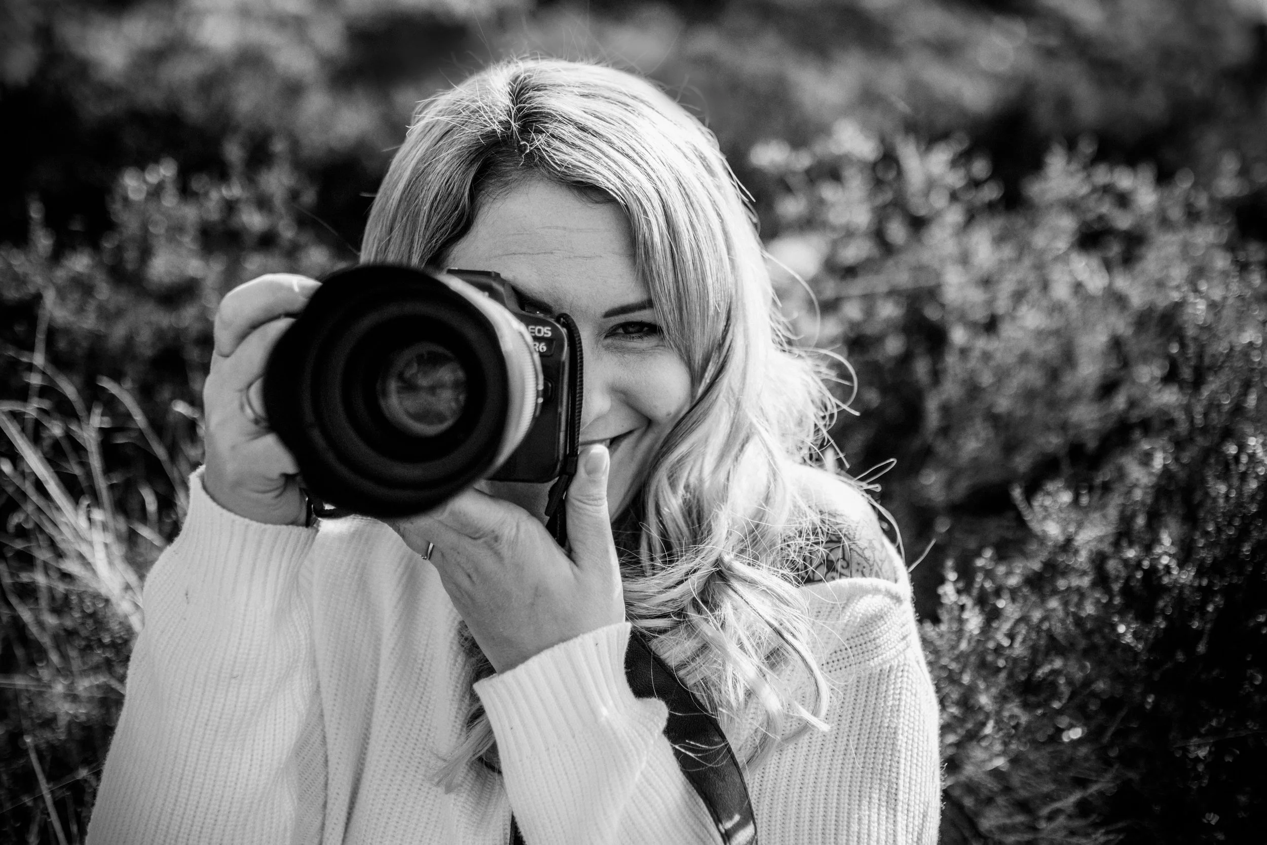 Frau mit langen Haaren hält eine Kamera in der Hand und fotografiert, während sie in die Kamera schaut und lächelt, im Freien mit Bokeh-Hintergrund.