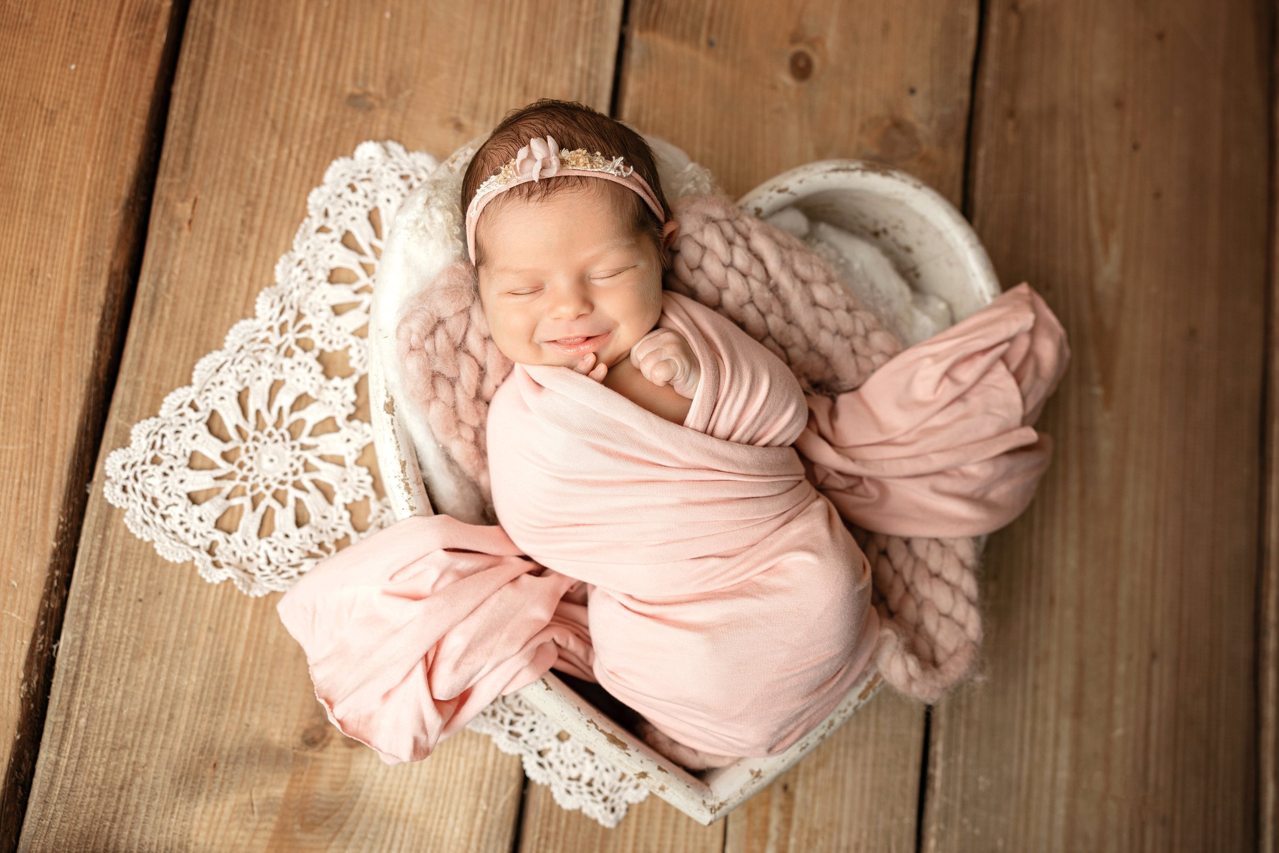 Süßes schlafendes Baby in einem rosa Tuch, mit Blumenhaarband, auf einem Herz aus Holz, umgeben von Spitze, auf einer Holzwand.