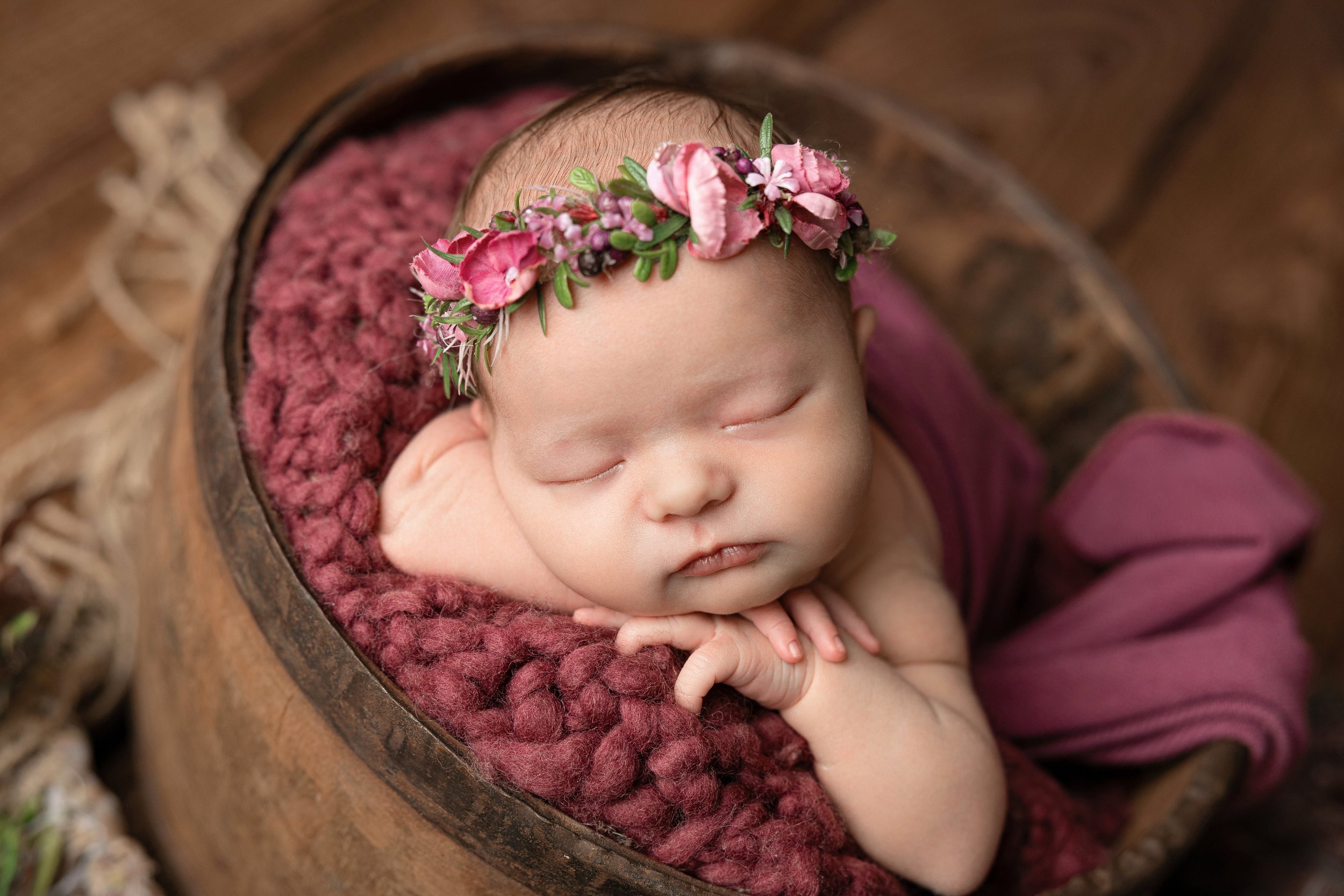 Ein schlafendes Baby mit einem Blumenkranz am Kopf liegt in einem Korb auf einem weichen, roten Kissen.