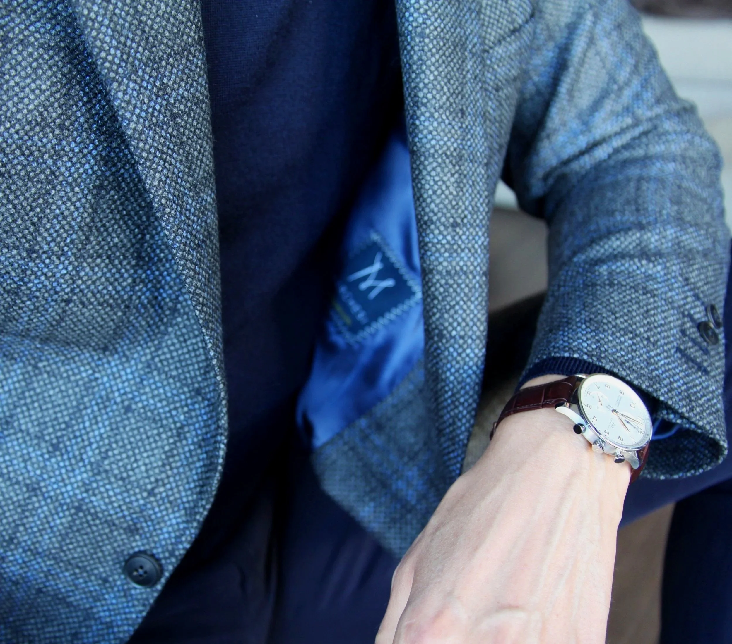 Close-up of a person's arm wearing a wristwatch, with the person dressed in a textured blue blazer and dark shirt, leaning on a surface with a blurred background.