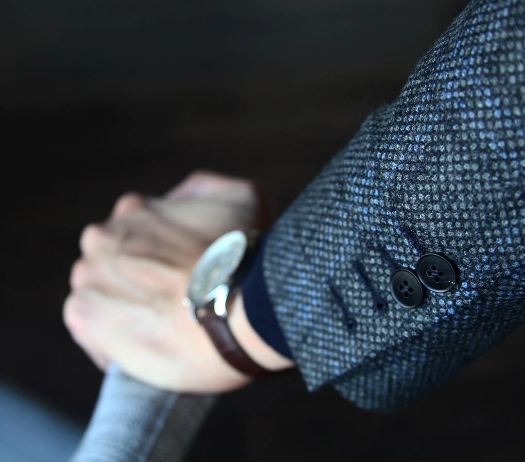 Close-up of a person wearing a textured gray blazer with black buttons, looking at their wristwatch, with a blurred background.