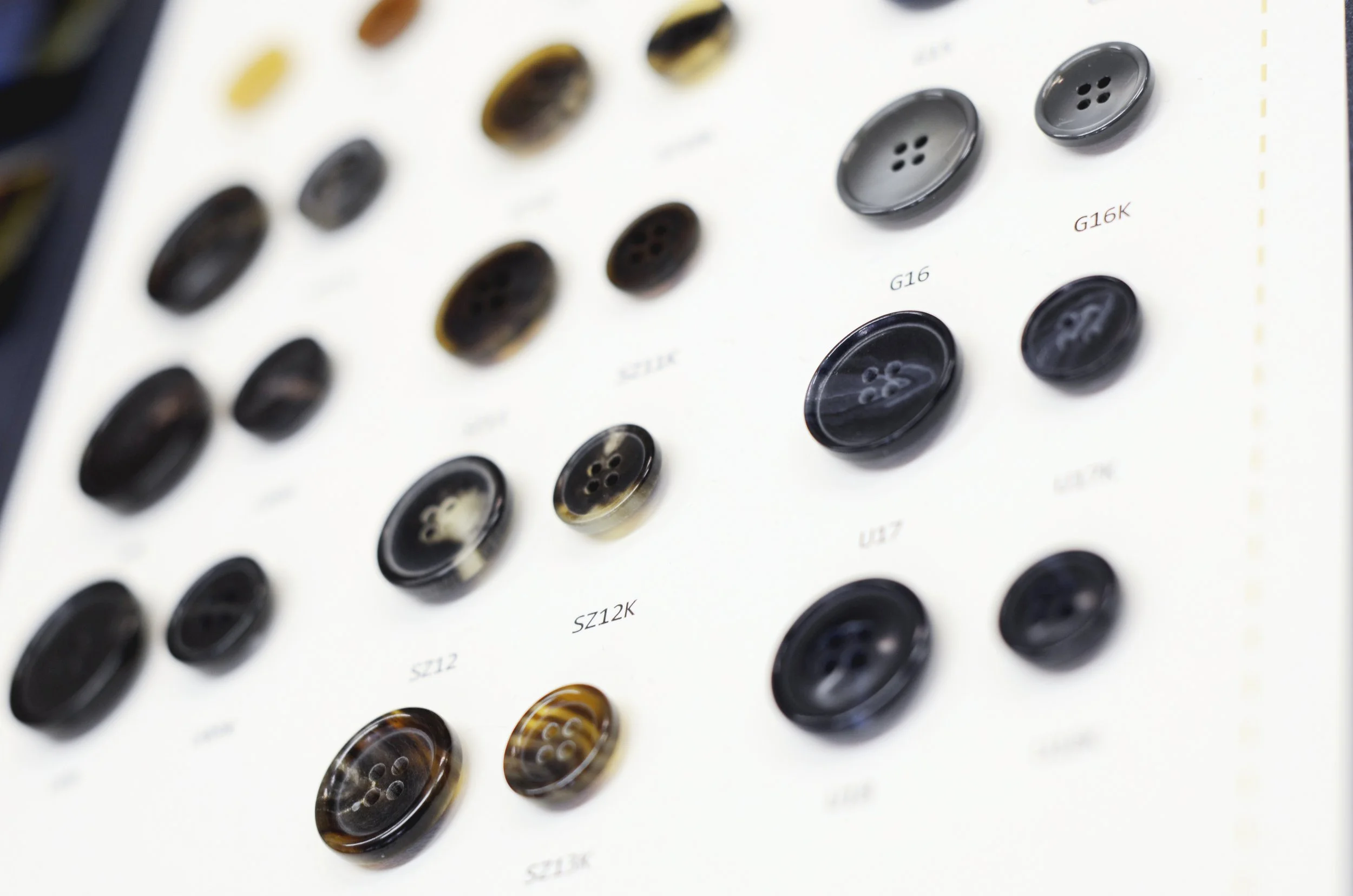 Close-up of a display board showing various black and brown buttons with four holes, labeled with different sizes and styles such as G16, G12K, and G16K.