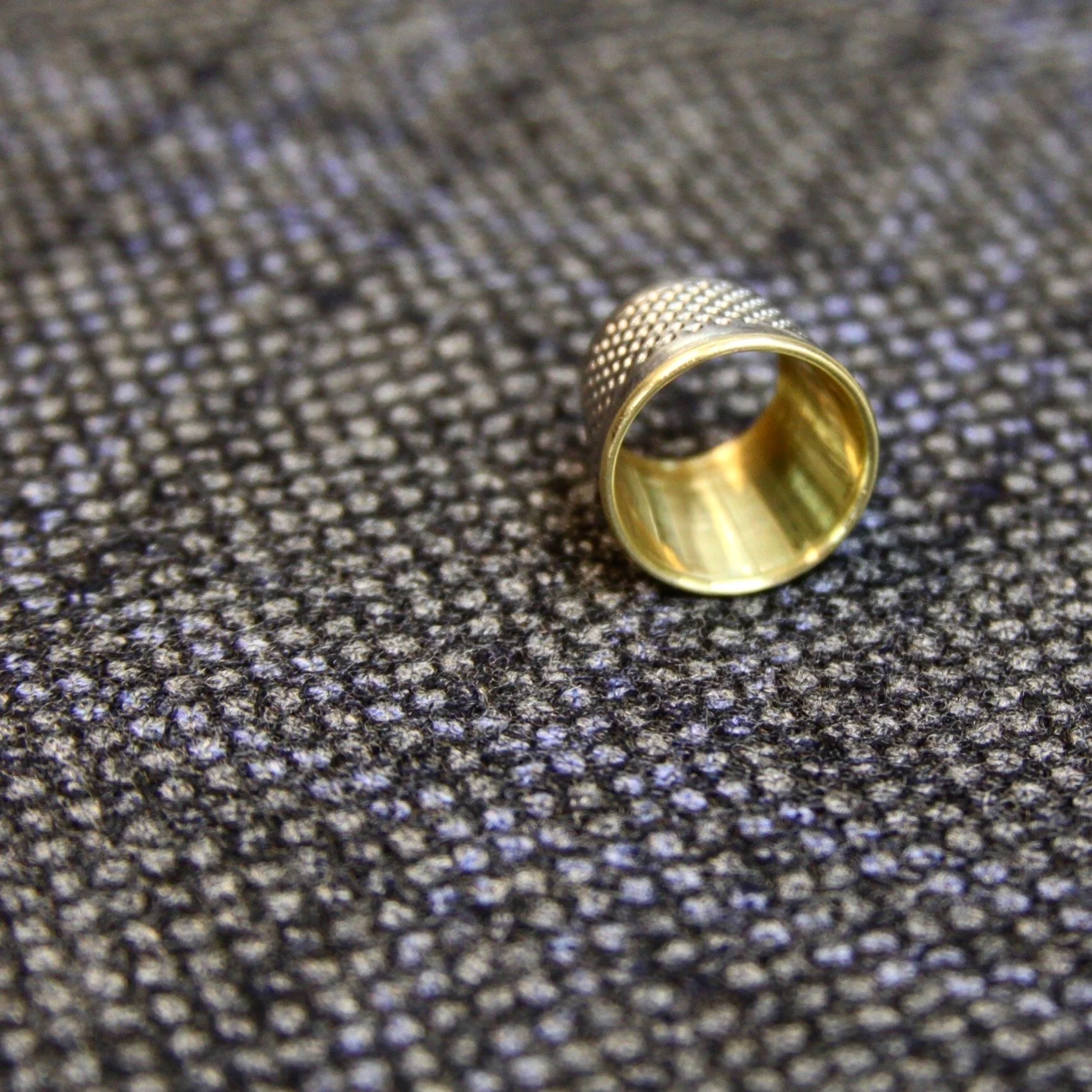 A metal thimble with a gold interior and textured exterior resting on a gray fabric surface.