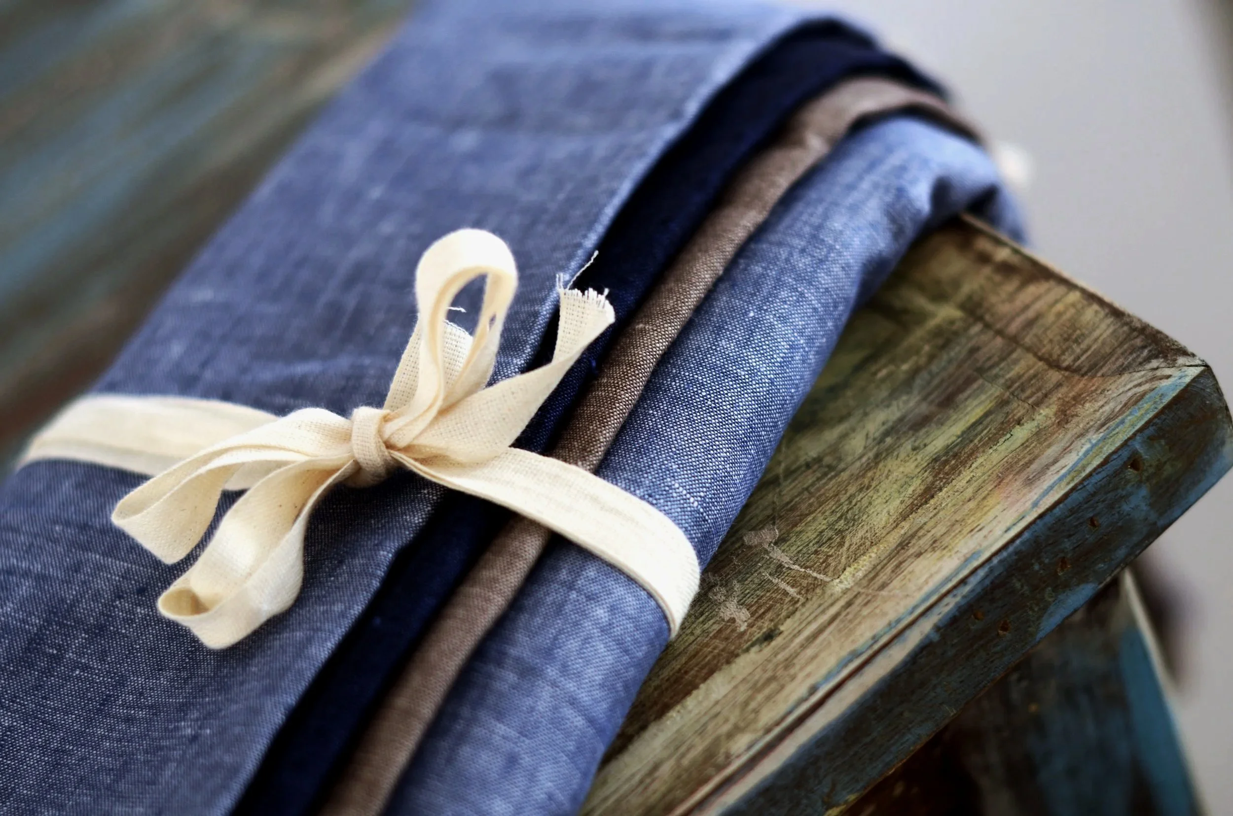 A close-up of a rolled stack of fabric in shades of blue and brown, tied with a cream-colored ribbon, resting on a wooden surface.