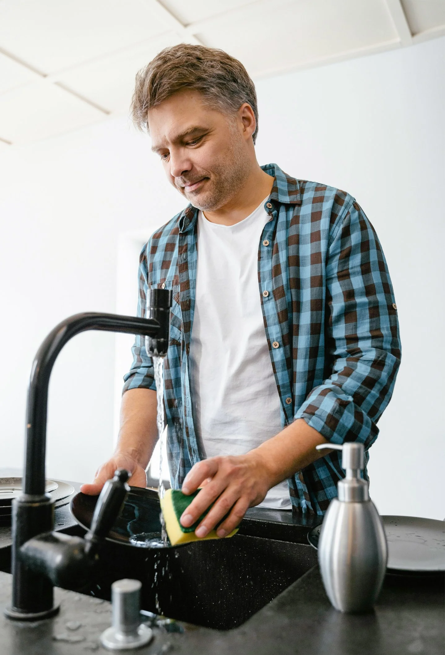 A man washing dishes at a kitchen sink, wearing a blue and brown checkered shirt and a white t-shirt underneath.
