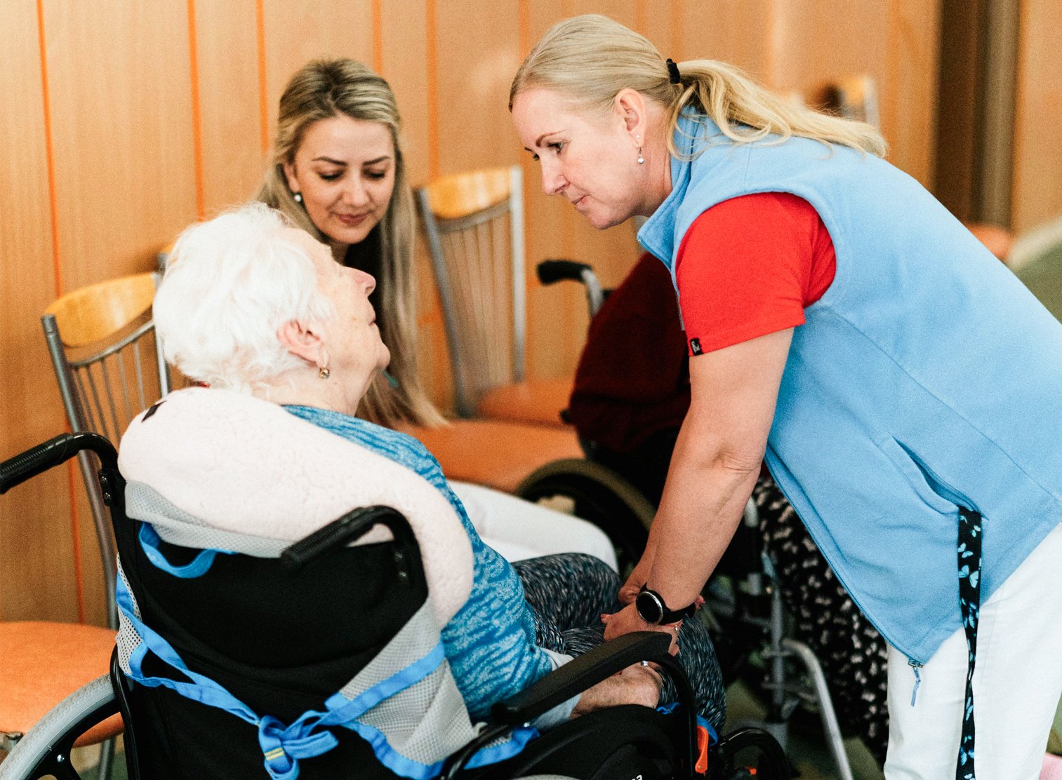 A healthcare worker and a visitor are attending to an elderly woman in a wheelchair in a healthcare facility. The healthcare worker, wearing a blue vest over a red shirt, is holding the woman's hand and leaning in to talk, while another woman with long blonde hair stands nearby, observing.
