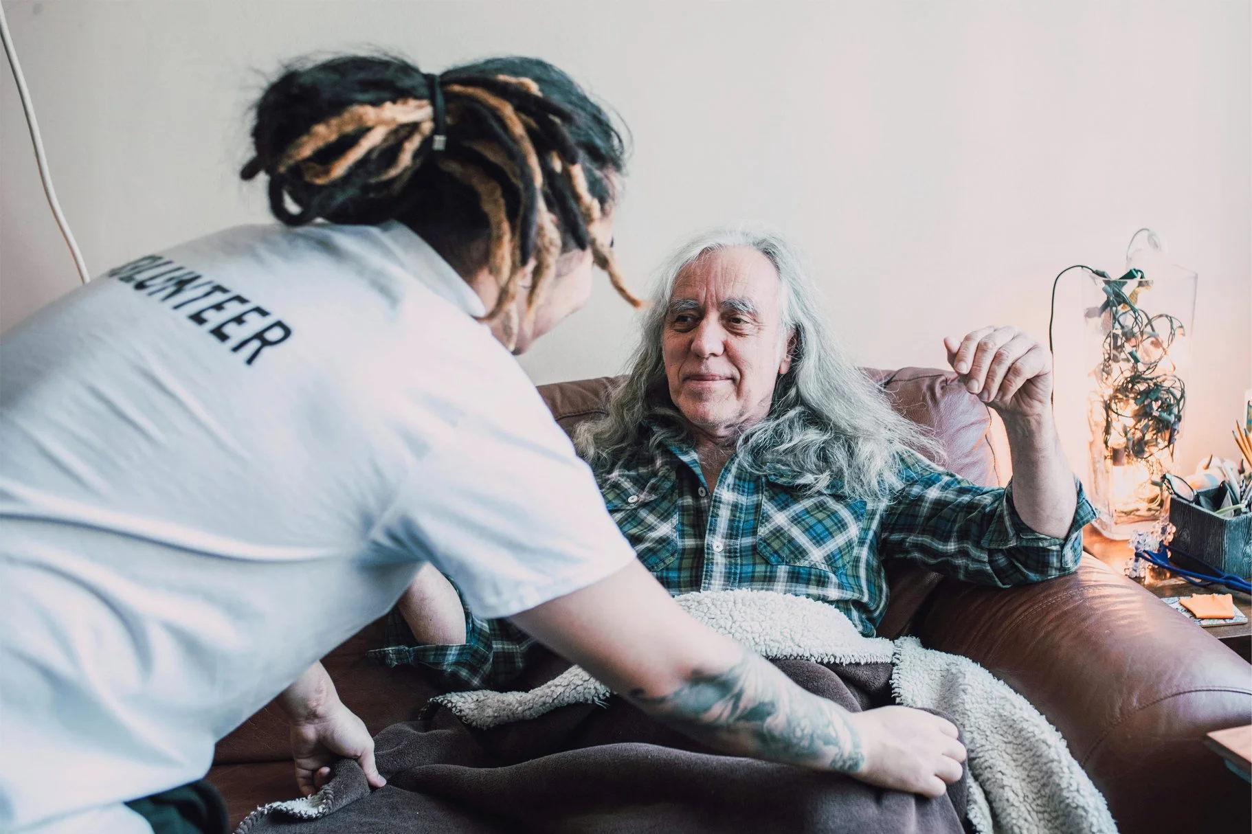 A young man with dreadlocks, wearing a white shirt that says 'COACHER' on the back, is helping an elderly man with long gray hair sitting on a couch in a living room. The elderly man is wearing a plaid shirt and is holding up his hand. There are plants and miscellaneous items on a table in the background.