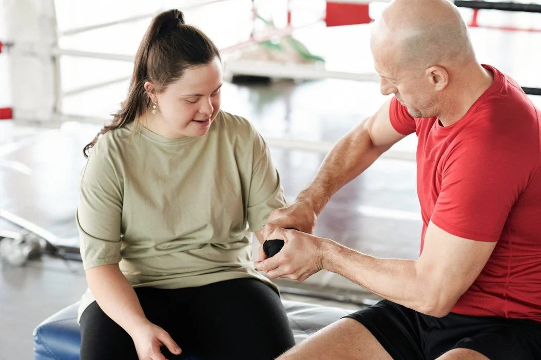 A young woman and a man are engaged in a weight-lifting training session, with the man assisting her with a dumbbell in a gym.