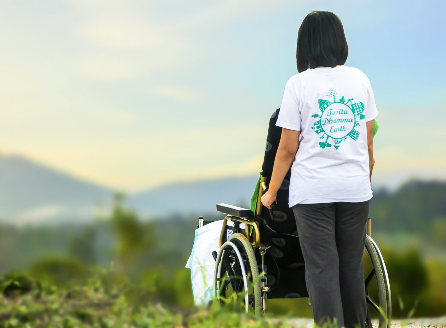 A person with shoulder-length dark hair stands outdoors with their back to the camera, wearing a white t-shirt that says 'Tavista Dharma Earth.' They are pushing a wheelchair with a black seat and large rear wheels through a grassy area. The background shows a landscape of distant hills under a partly cloudy sky.