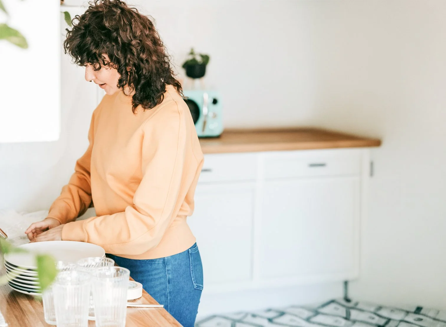 A woman with curly dark hair wearing a peach long-sleeve shirt and jeans is standing in a kitchen, preparing food at the counter with stacked plates and glasses nearby.