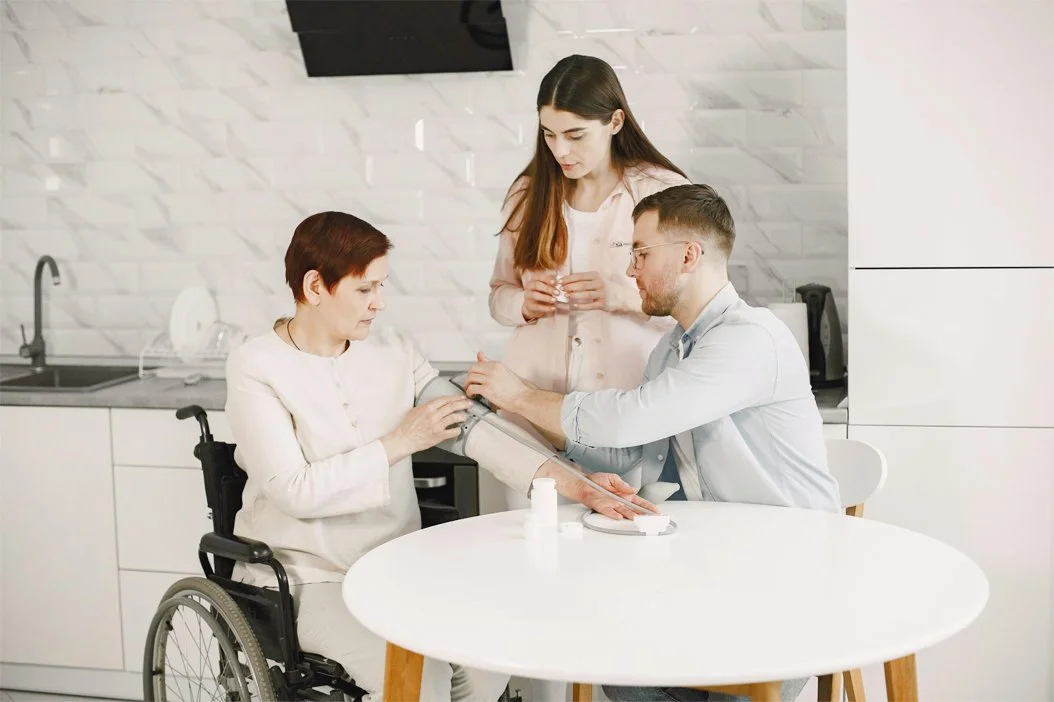 A healthcare worker in a white coat checks a woman's blood pressure while another woman observes in a modern kitchen or clinic setting.