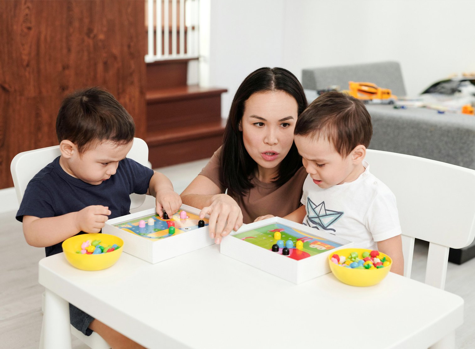 Woman playing a board game with two young boys at a white table in a living room.