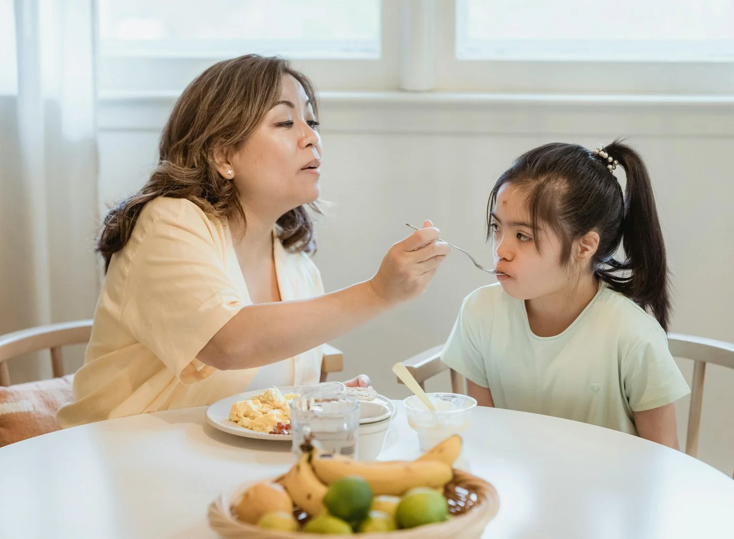 A woman feeding a young girl breakfast at a dining table, with a bowl of cereal, toast, eggs, and a basket of fruit in the foreground.