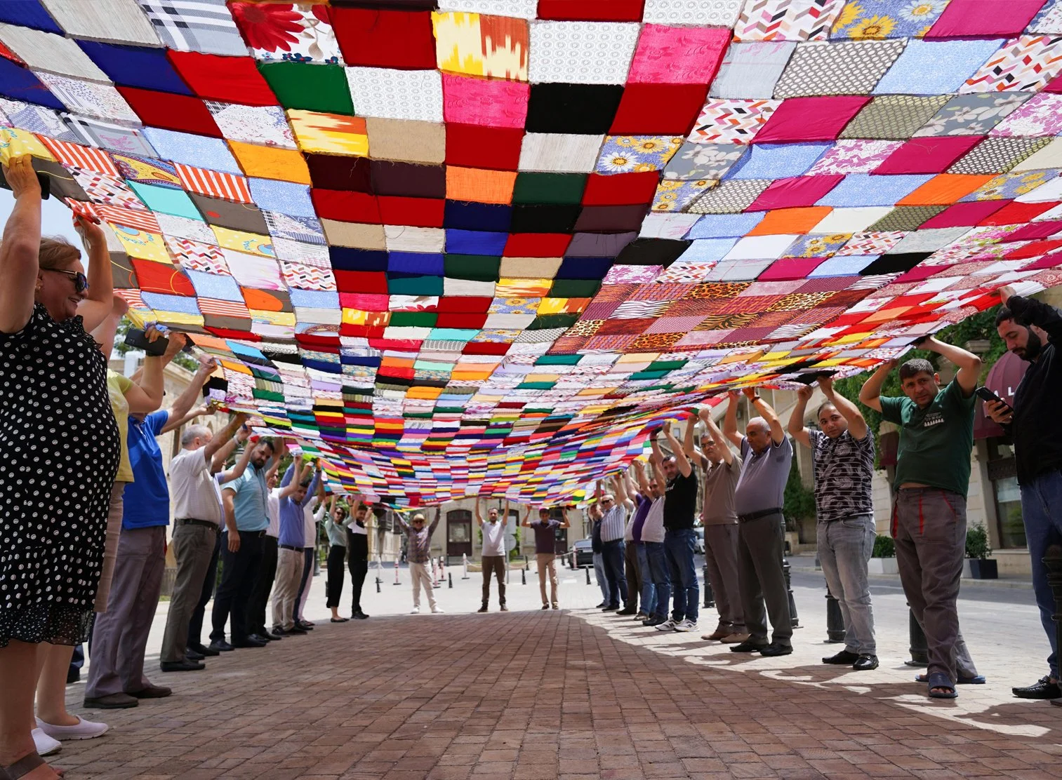 People standing in a circle holding up a large, colorful quilt canopy made of many fabric squares overhead in an outdoor urban setting.