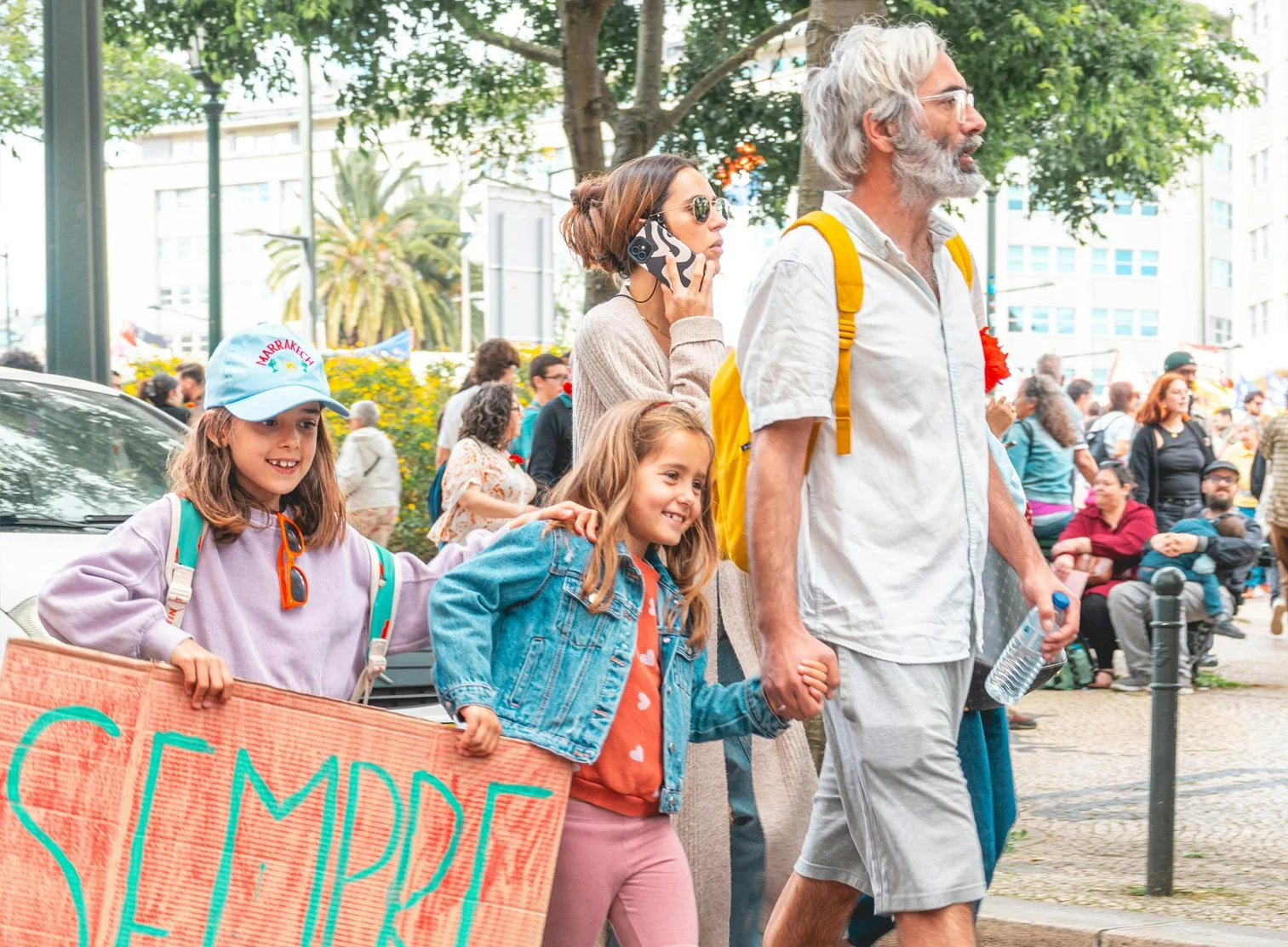 People participating in a protest or demonstration walking on a city street. Two young girls carrying a large orange banner, one wearing a light purple hoodie and a blue cap, the other in a denim jacket. An older man with gray hair and a beard holding a water bottle, wearing a white shirt and gray shorts, walking beside a woman talking on a cellphone, wearing sunglasses and a beige sweater. Many other people are in the background sitting and standing, gathered around in an urban park setting.