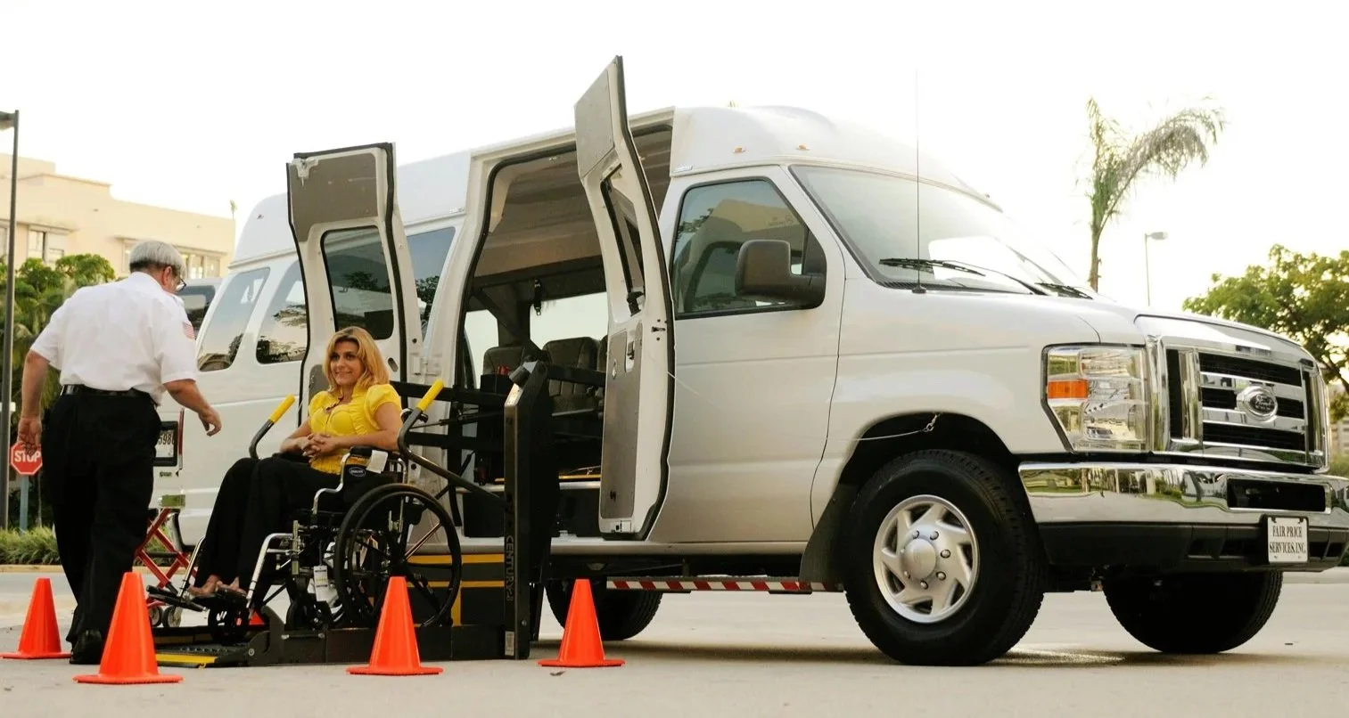 A woman in a wheelchair is being assisted by a man near a white van with an accessible lift, in an outdoor setting.