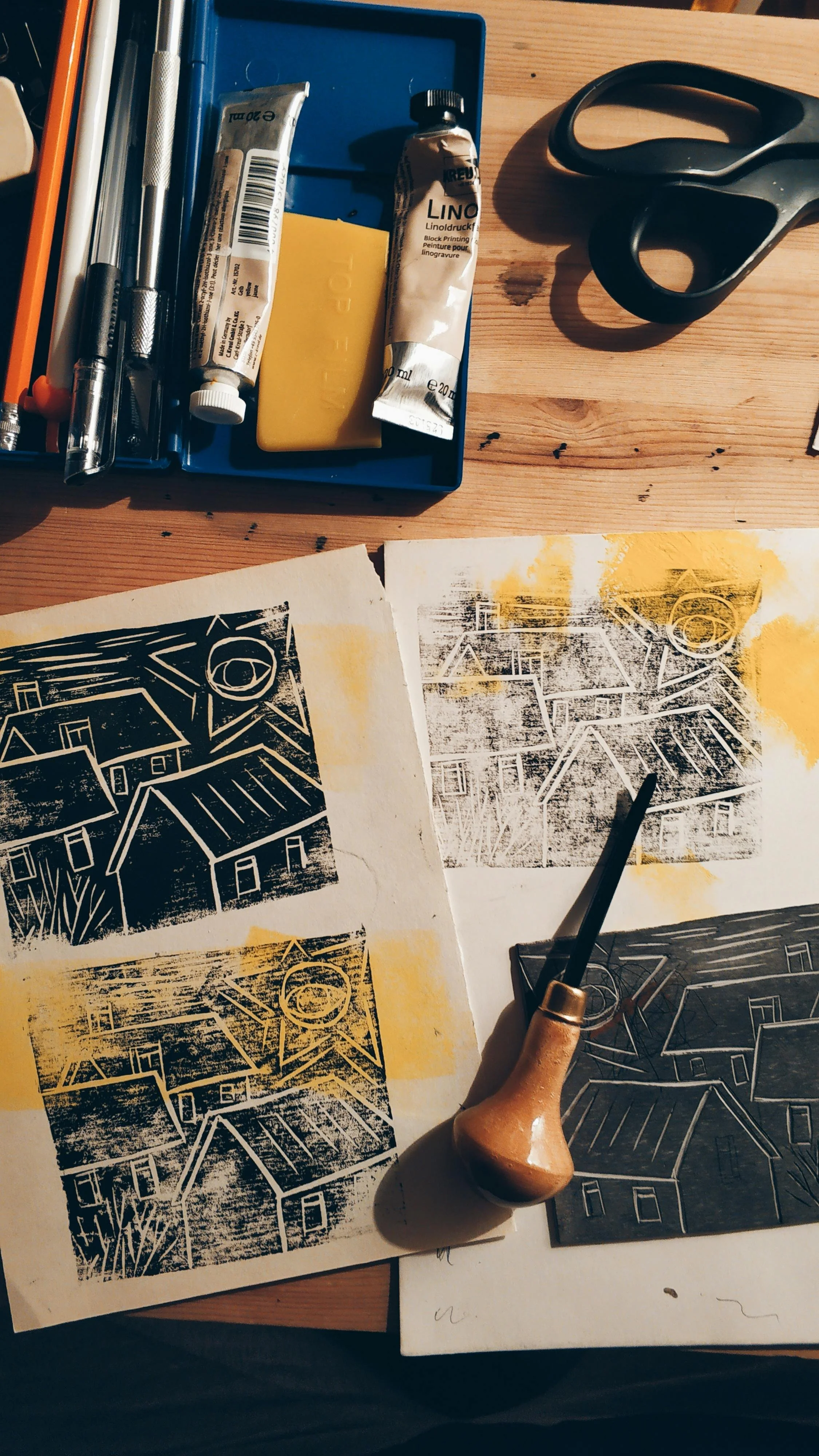 A printmaking or block printing workspace with a black-and-white house design, a carving tool, and printing blocks on a wooden table. Art supplies such as a tube of linoleum ink, scissors, and metal tools are also visible.