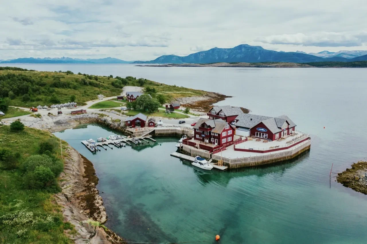 Landskap med sjø, fjell i bakgrunnen og en liten havn med båter og røde bygninger