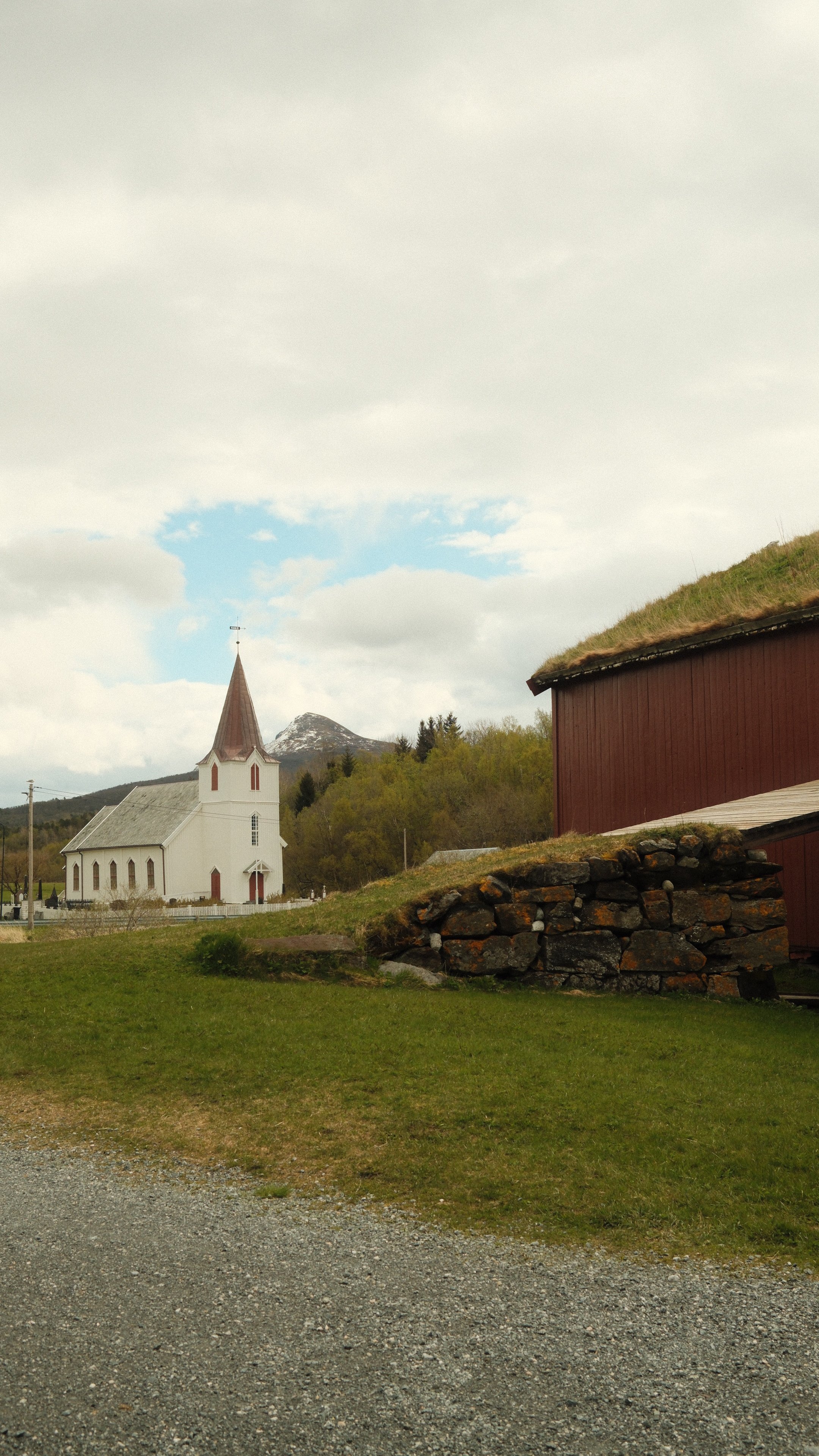 En landsby med en hvit kirke med spiss tårn, omgitt av grønne åser og fjell i bakgrunnen, under en overskyet himmel.