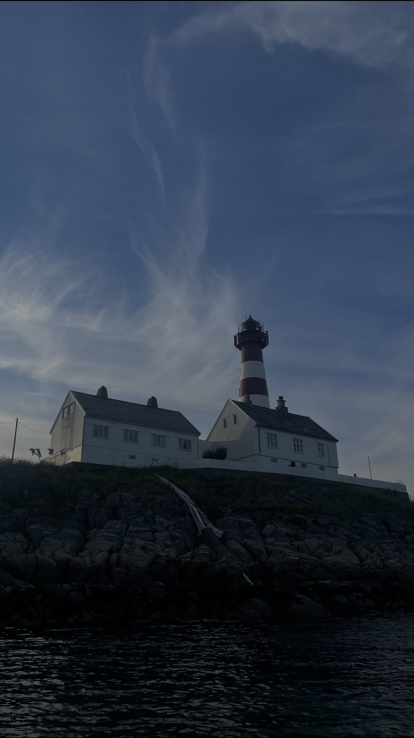 En fyrtårn og to hvite hus ligger på en klippe ved vannet, med en klar blå himmel og strøk av skyer i bakgrunnen.