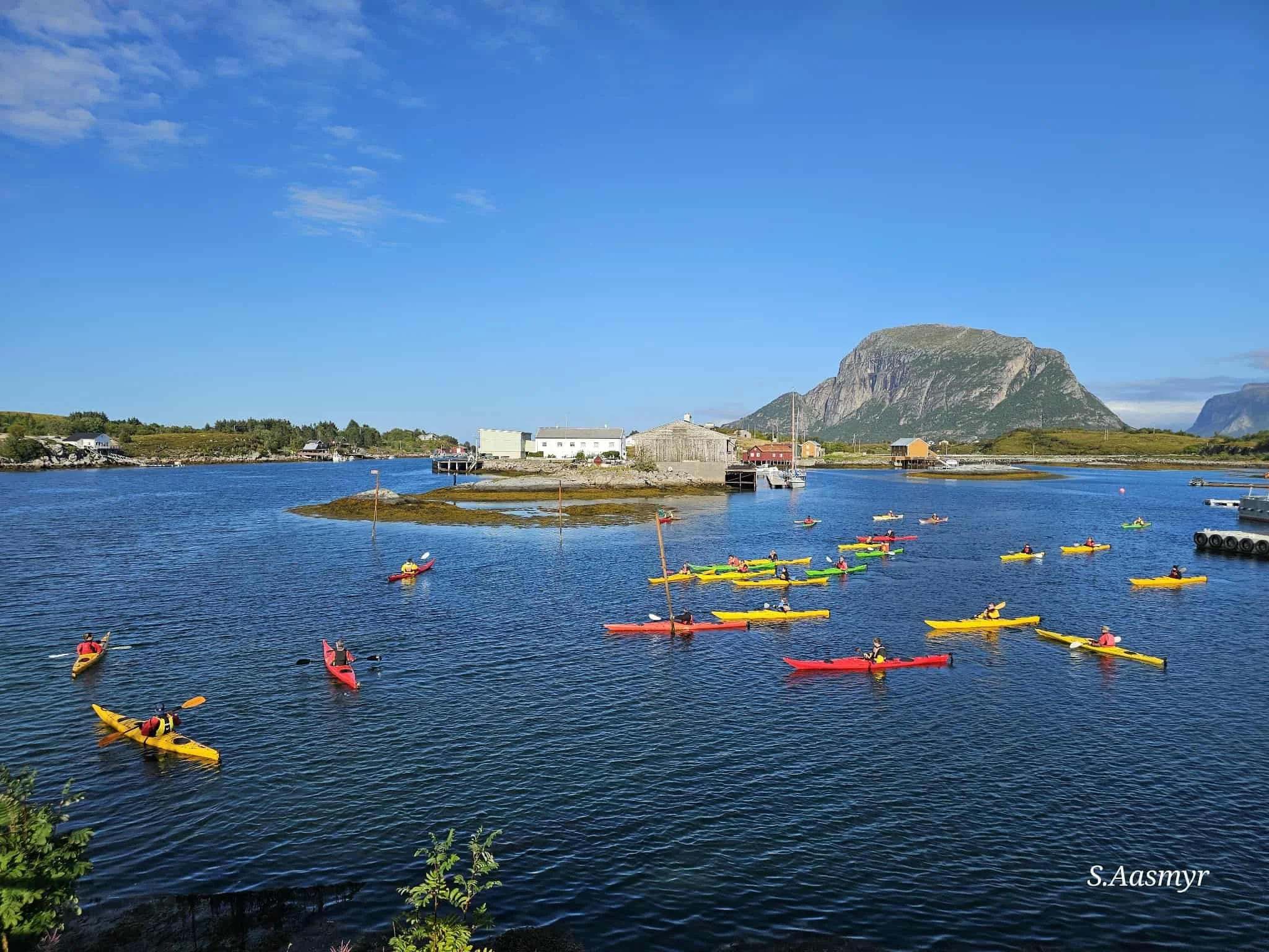 Fjell, vann og små hus ved sjøen, med flere folk i kajakker på vannet under en klar blå himmel.