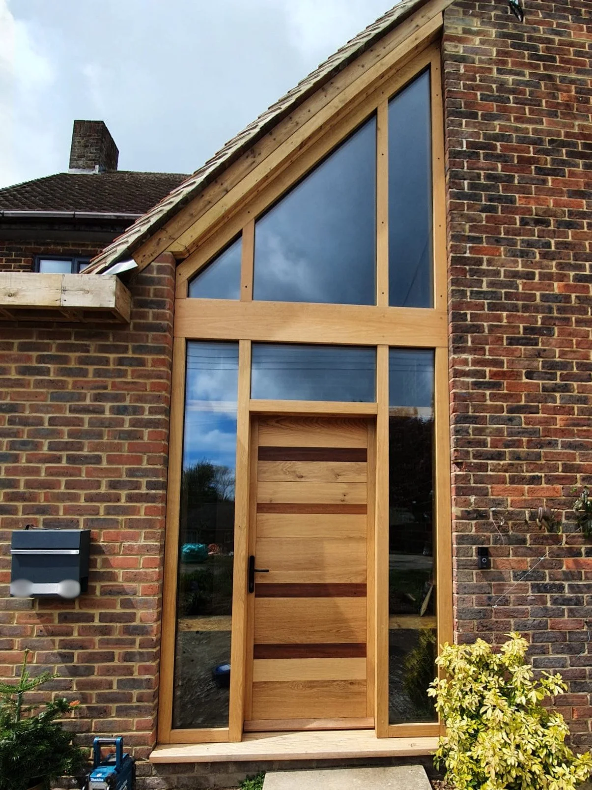 The front of a brick house with a newly built wooden door and large glass windows above and beside it. The house has a triangular roof with a section under construction on the left side. A mailbox is attached to the brick wall on the left, with some 