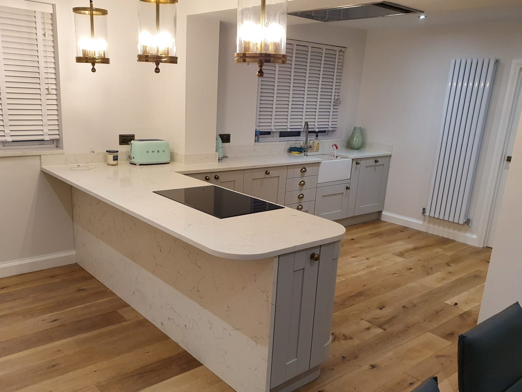 Modern kitchen with white cabinets, a white marble island, a black induction cooktop, and wooden floors. Hanging above are three gold and glass light fixtures. There are two windows with white blinds, a radiator on the wall, and some kitchen items on