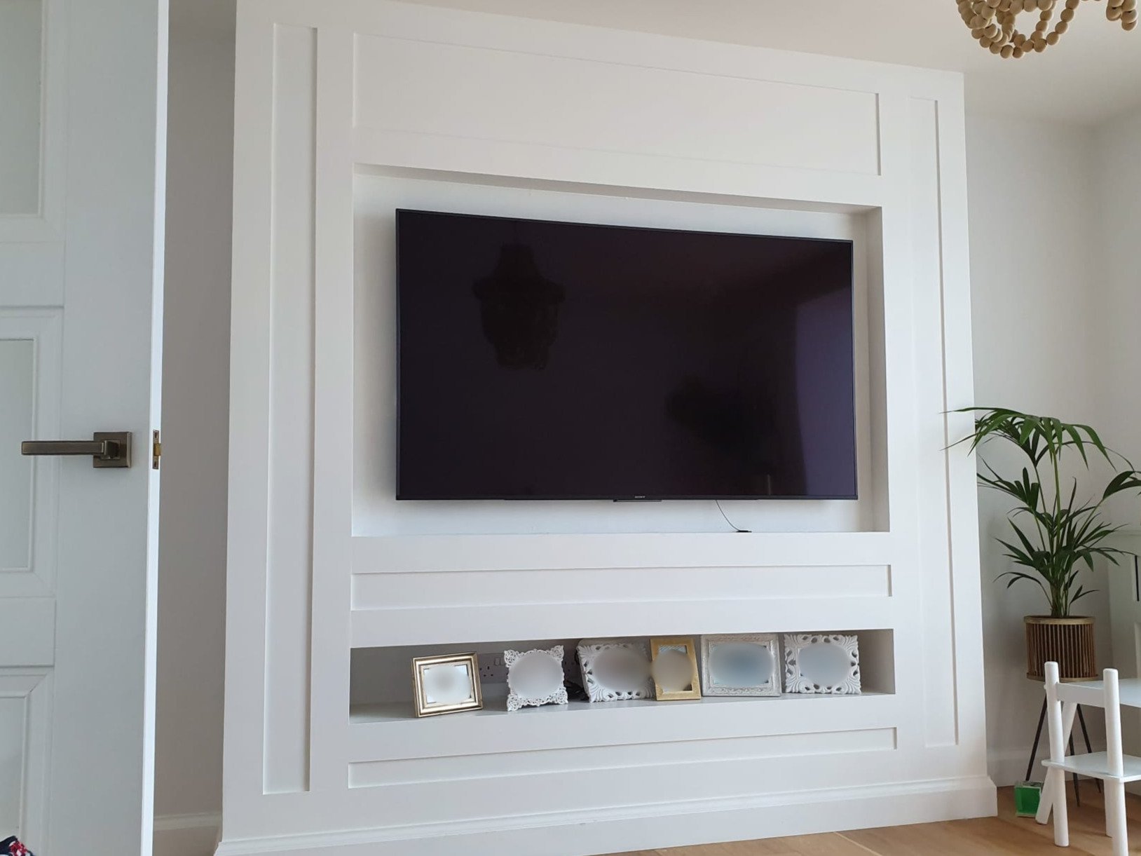 Living room with a mounted flat-screen TV on a white built-in wall and a potted plant to the right.