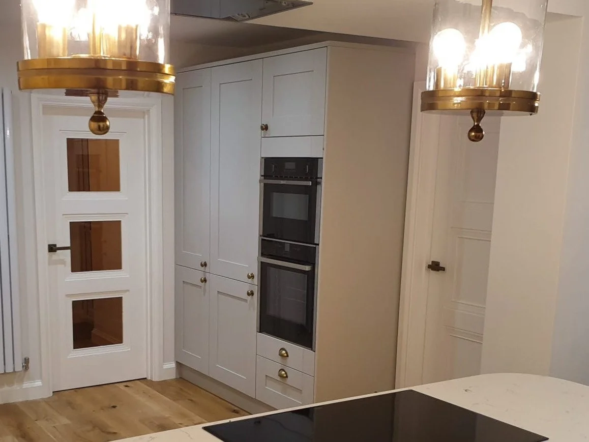 A kitchen with white cabinets, black double ovens, a white door with glass panels, hardwood floors, and two gold and glass pendant lights.
