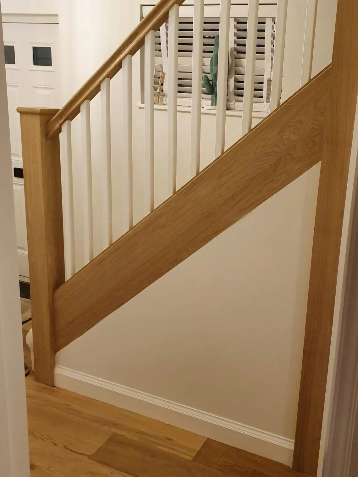 Part of an interior staircase with a wooden handrail and white vertical balusters, with a window with shutters and decorative cactus plant in the background.