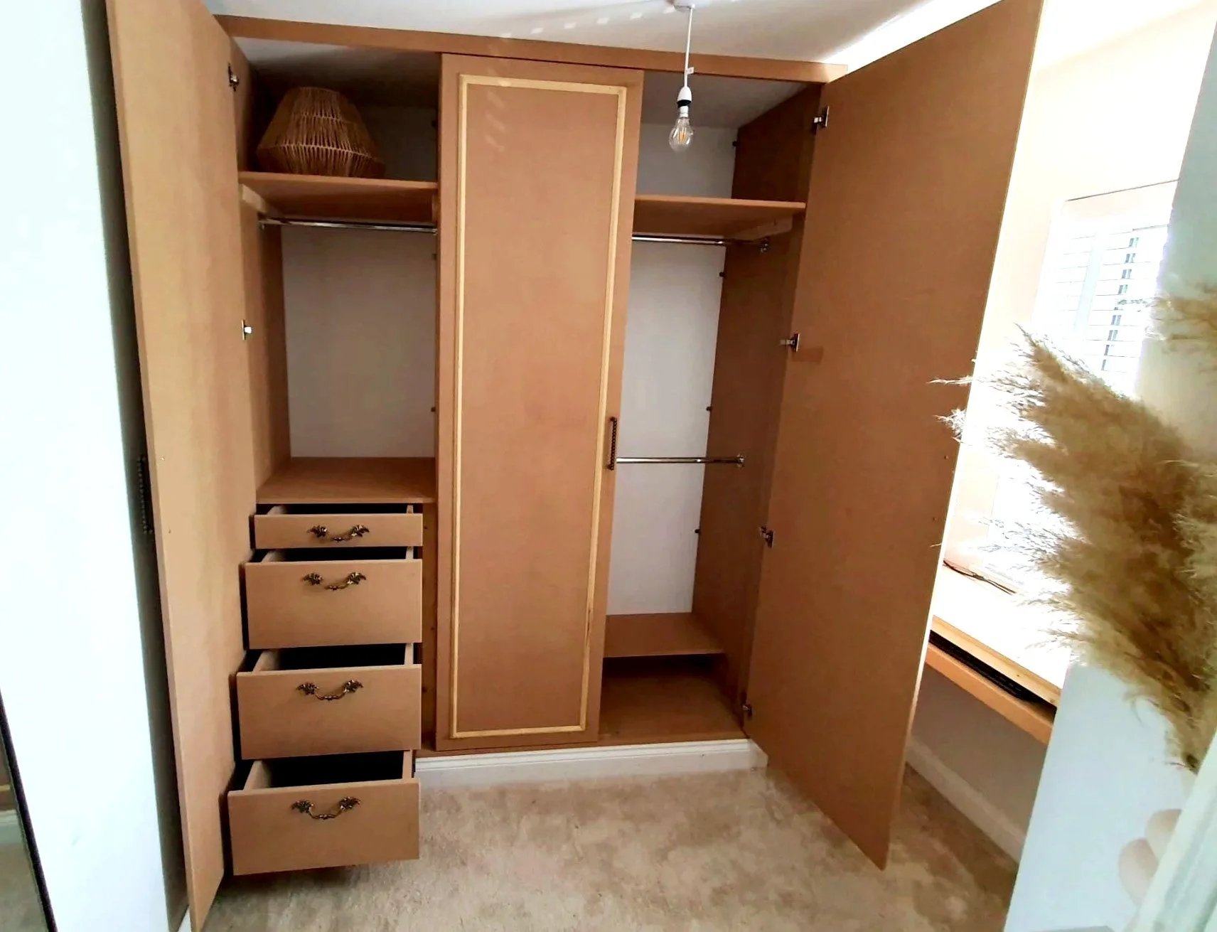 Open wooden closet with drawers on the left, hanging space for clothes in the middle, and a built-in cabinet with a door above. A exposed light bulb hangs from the ceiling, and a window with white shutters is on the right, letting in natural light.