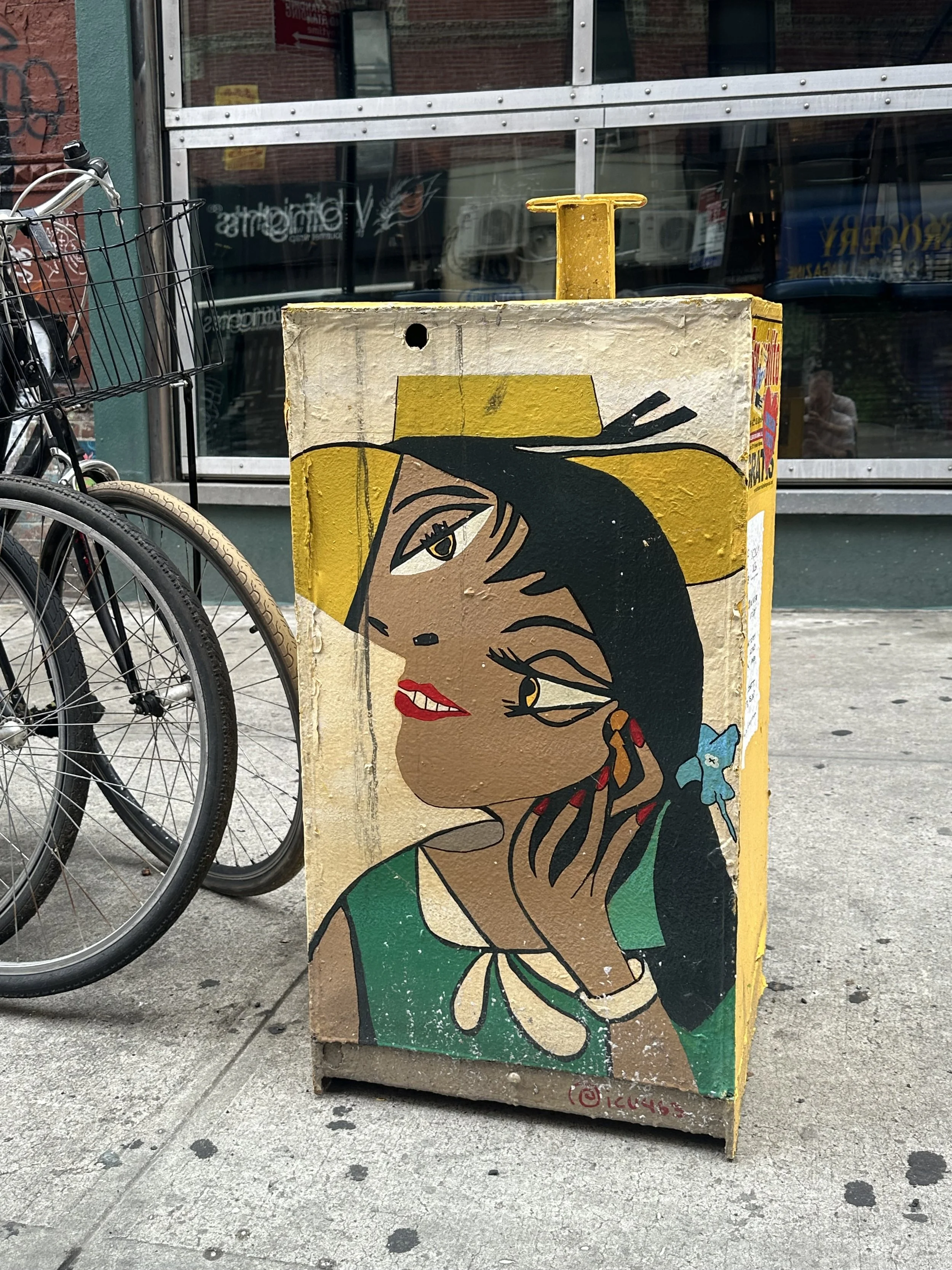 Colorful street-art portrait on a New York sidewalk beside parked bicycles and a storefront window.