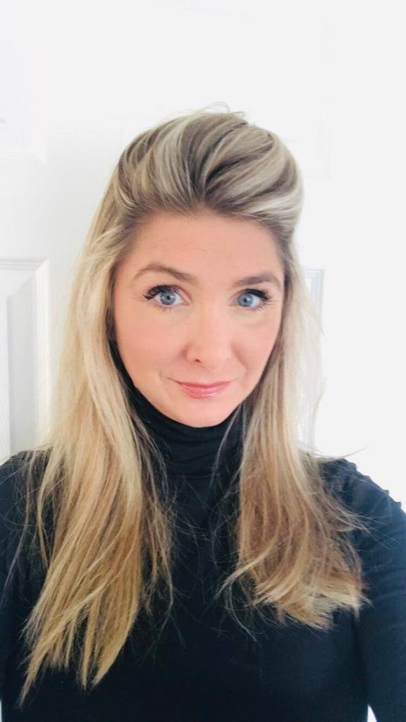 A woman with long blonde hair and blue eyes wearing a black turtleneck, standing indoors against a white wall.