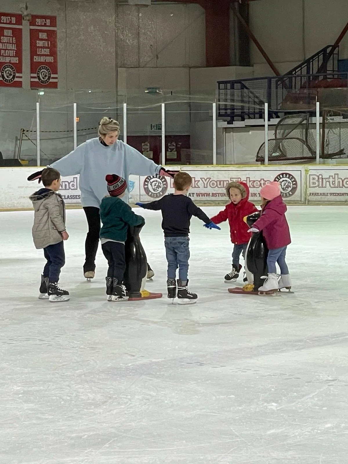 A group of young children in ice skates gathered in a circle on an ice rink, listening to a female instructor in a white sweater, who is explaining or demonstrating skating techniques. The children are wearing winter jackets and hats, and are standing near penguin-shaped skating aids.