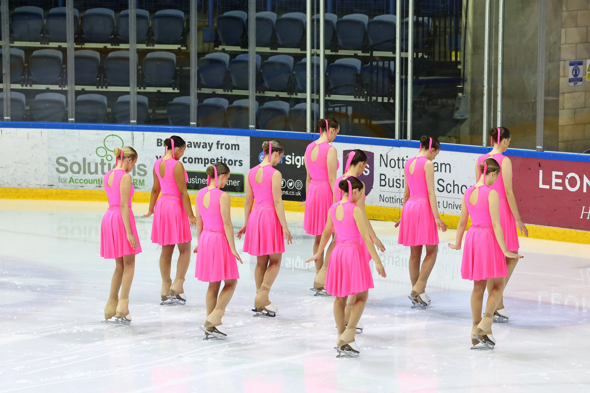 A group of figure skaters dressed in matching bright pink costumes practicing on an ice rink. Behind them, empty blue spectator chairs and advertising boards are visible.