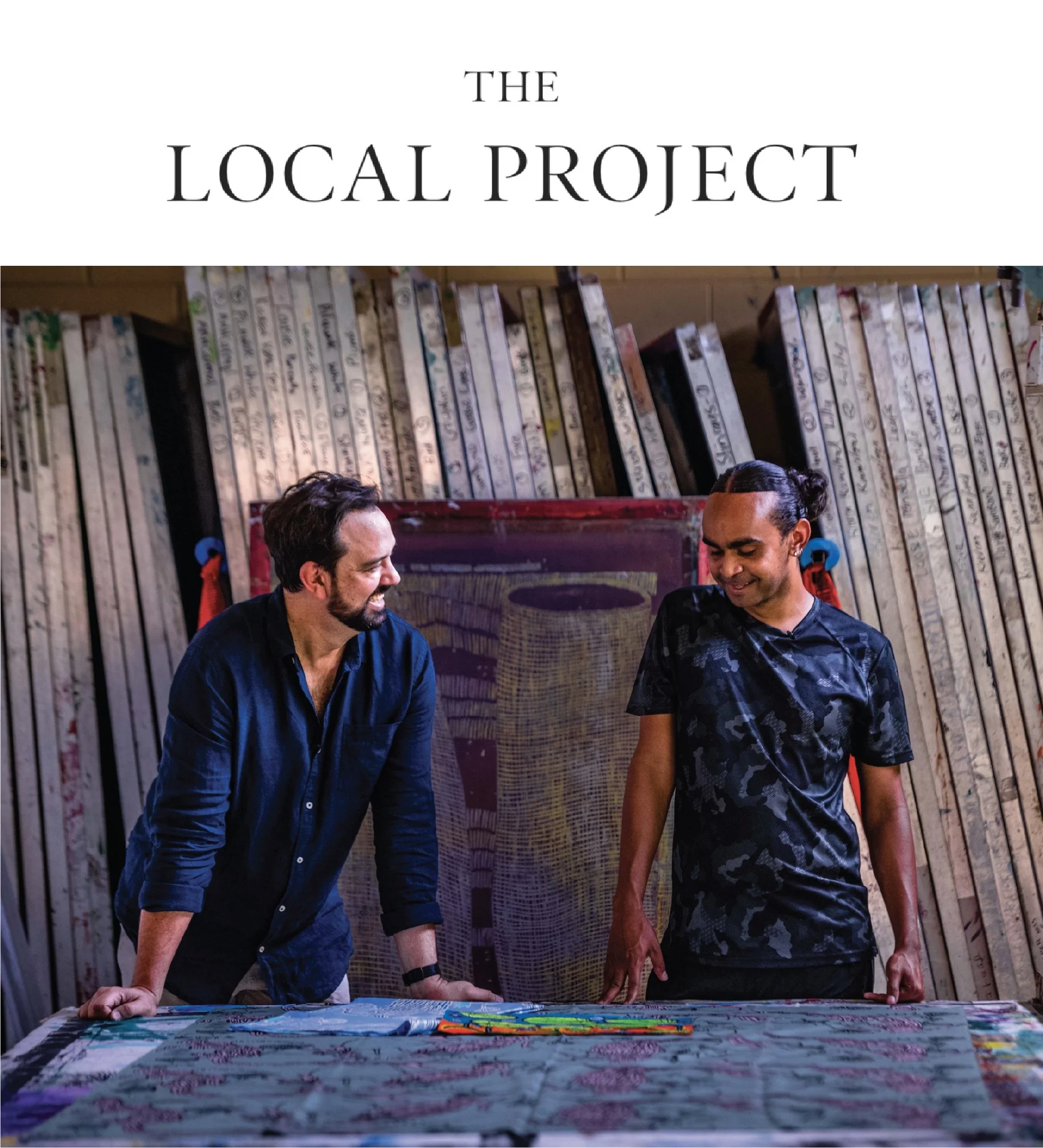 Two men standing behind a table covered with colorful printed fabrics, smiling and engaging in conversation in a room with a wooden wall decorated with vertical painted planks.