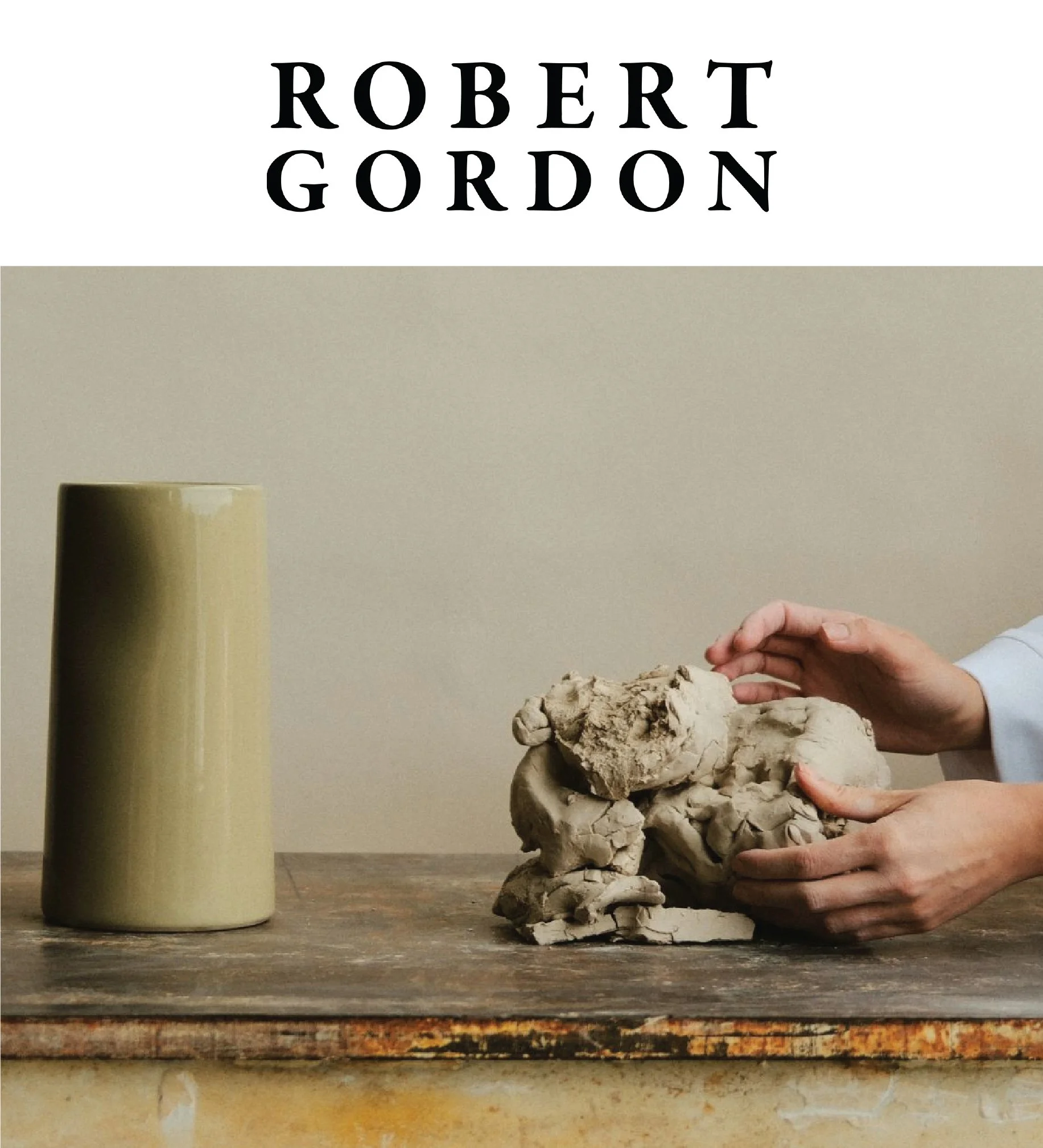 A person's hands shaping a large piece of clay on a wooden table, with a cream-colored ceramic vase in the background and a cream-colored wall.