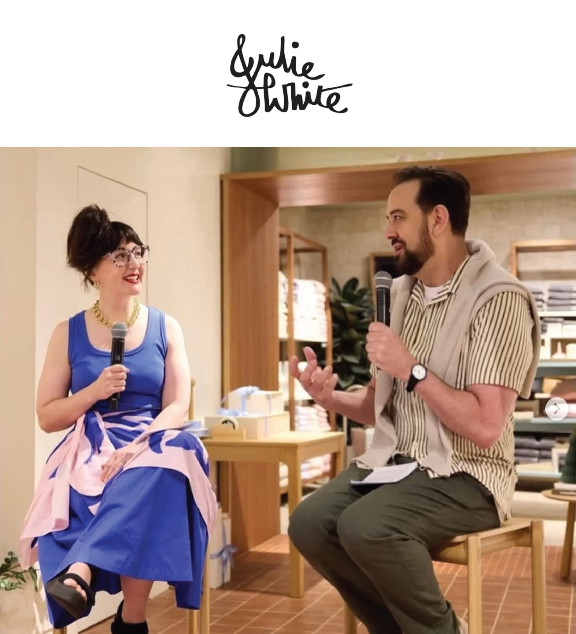 Two people sitting on a wooden bench indoors, engaging in conversation. The woman on the left is wearing a blue dress, glasses, and a gold necklace, while the man on the right is dressed in a striped shirt and suspenders, holding a microphone. They are facing each other in a casual setting with shelves and plants in the background.