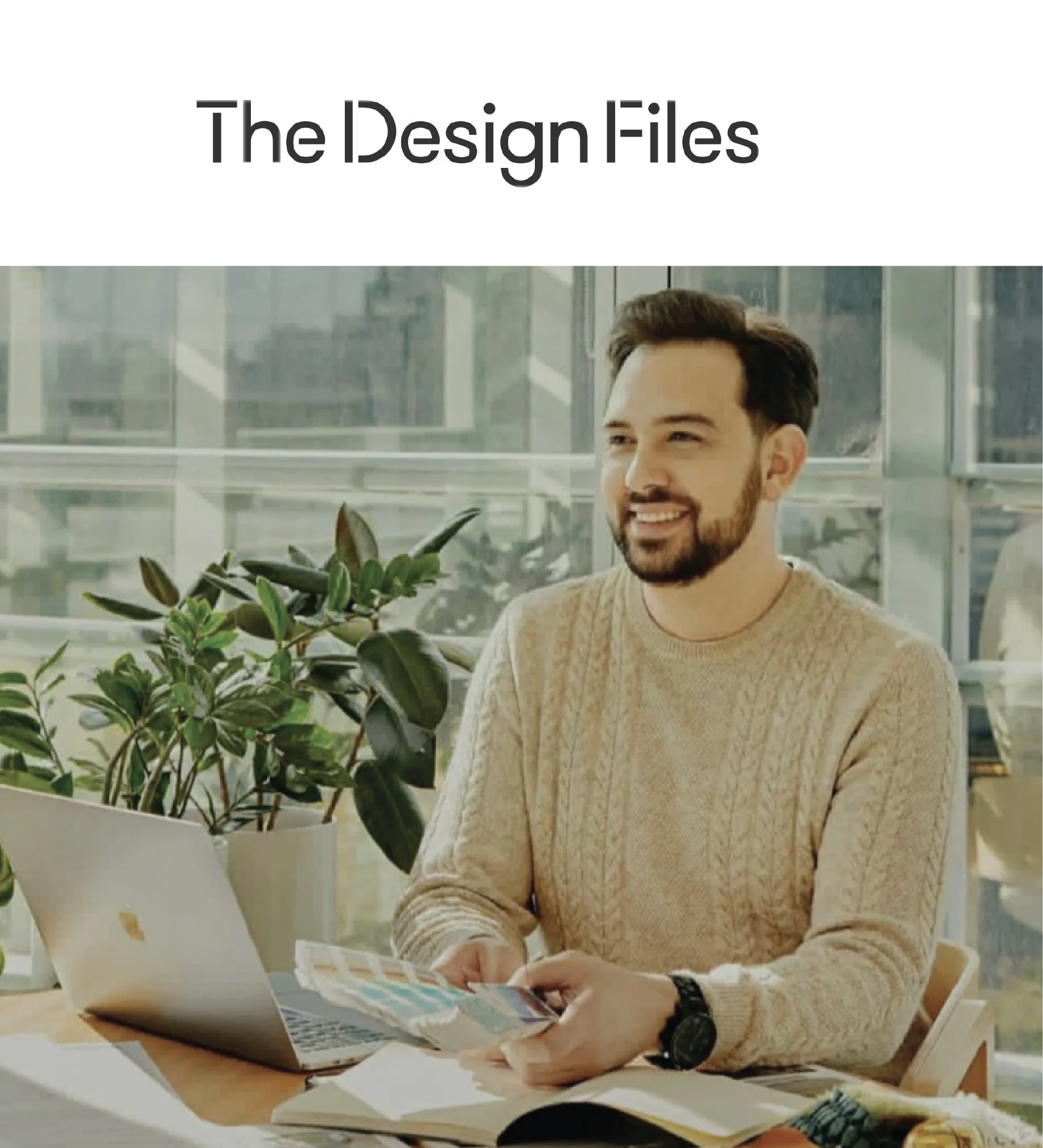 A man sitting at a table looking at a laptop and a magazine, with a potted plant nearby, in a bright modern office with large windows.