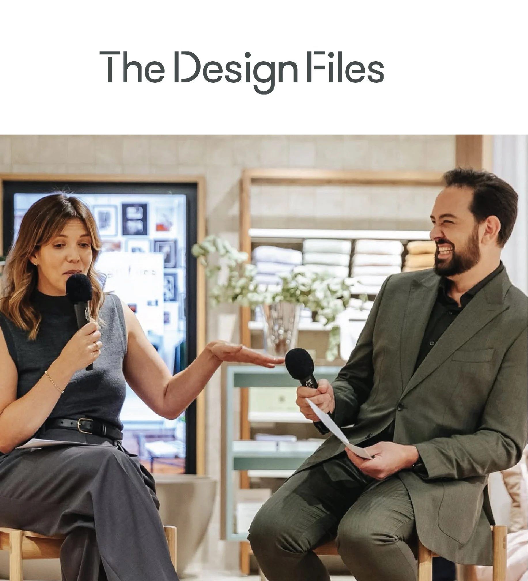 Two people seated and talking in a room with wooden furniture. The woman on the left is holding a microphone, and the man on the right is gesturing with his right hand while holding a paper. Both are smiling.
