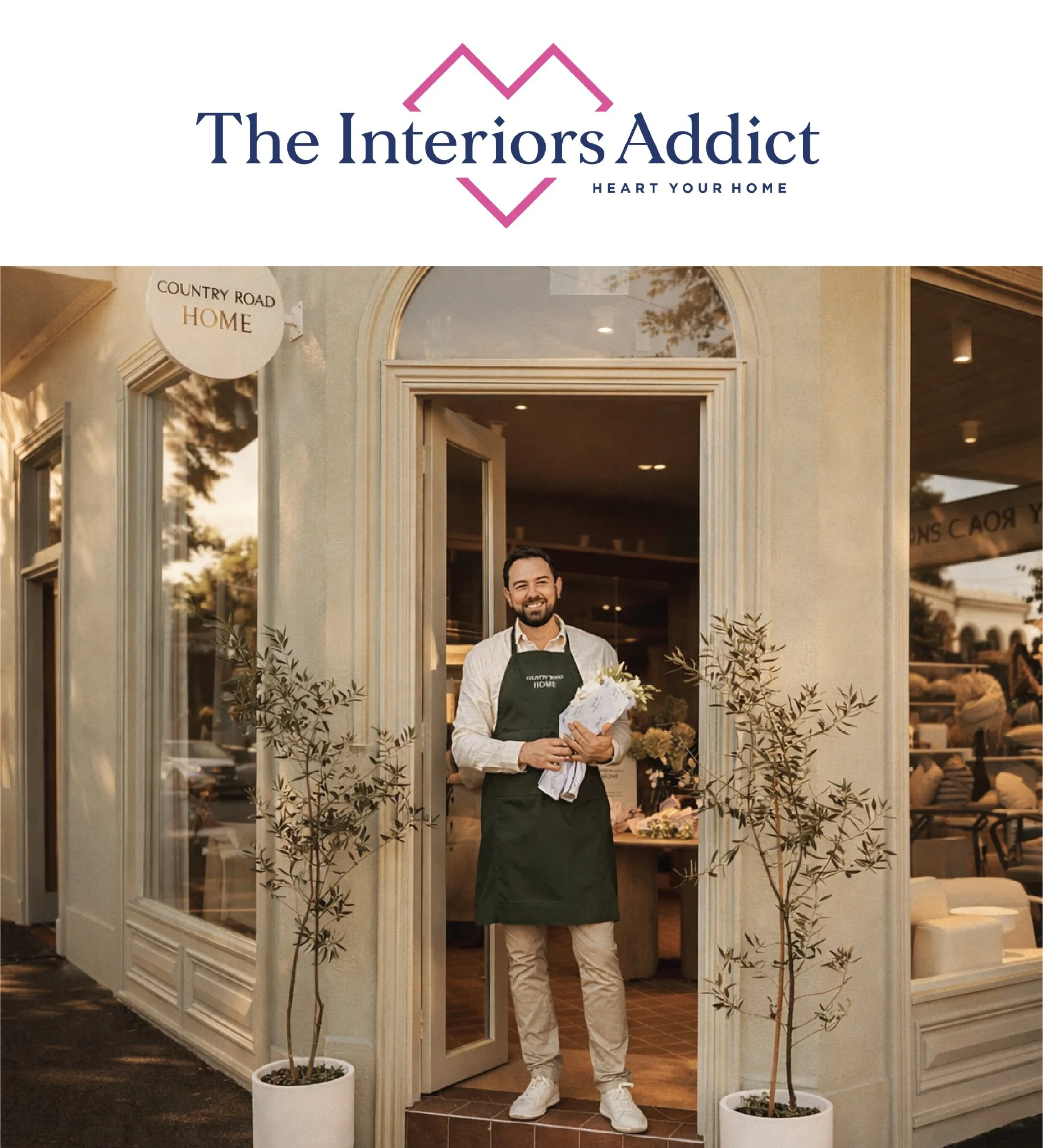 A man standing at the entrance of a shop, holding a bouquet of flowers, smiling, wearing a white shirt, beige pants, white shoes, and dark green apron, with potted trees on either side of the doorway.