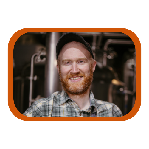 A man with a beard and a black cap smiling in an industrial setting with pipes and machinery in the background.