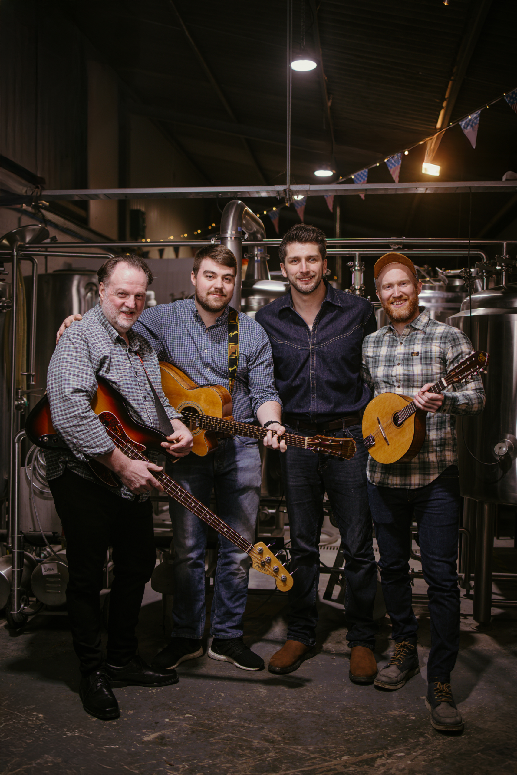 Four men standing together in an industrial setting, with three of them holding guitars, smiling at the camera. The background features brewing equipment and decorative bunting with American flags, indicating a festive, musical atmosphere.