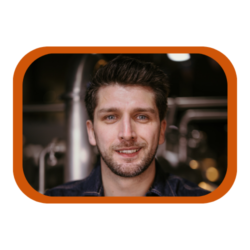 A man with brown hair and a beard smiling at the camera in a kitchen or restaurant setting.