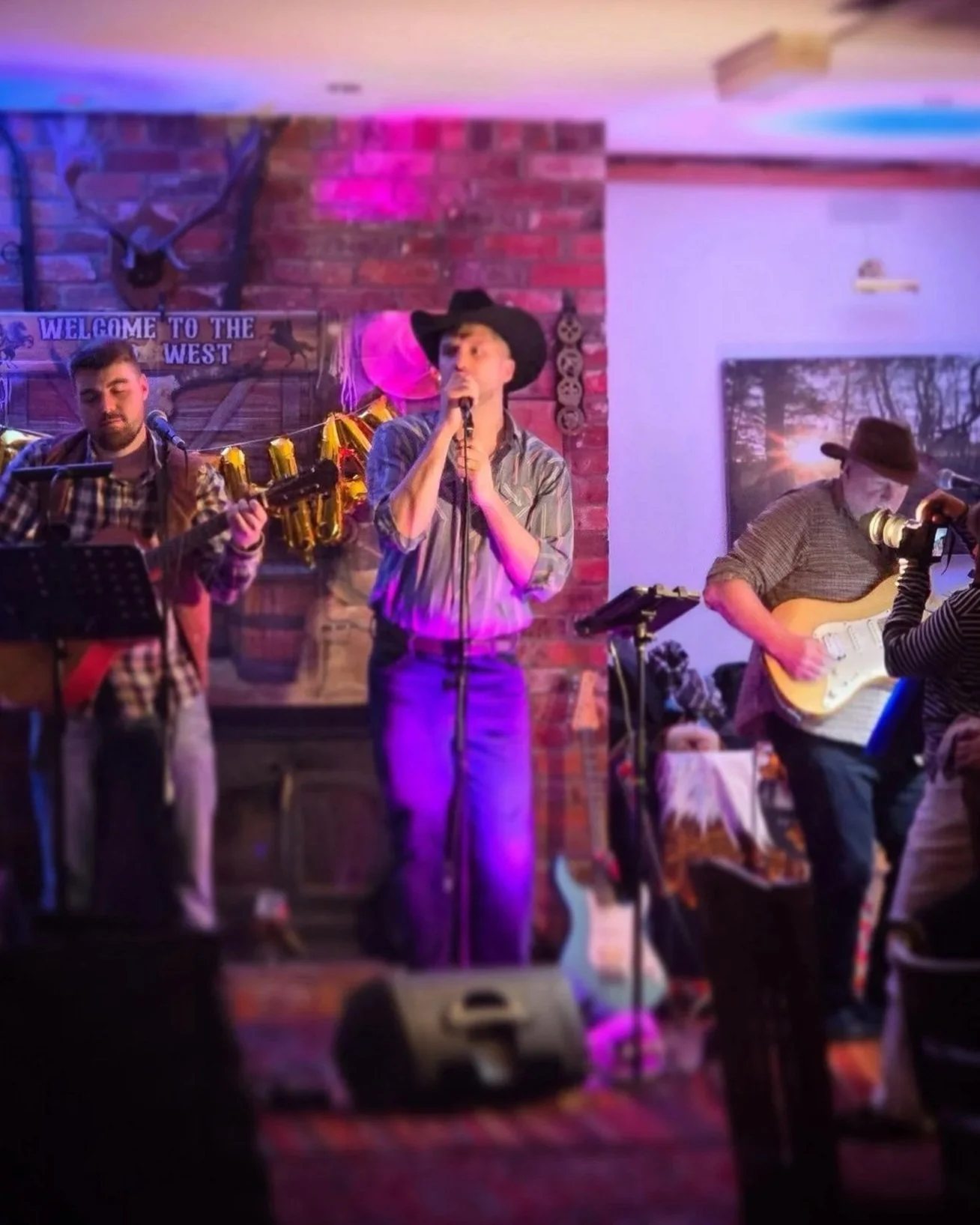 A group of musicians performing on a stage with a brick wall background and a 'Welcome to the West' sign. The performers include a woman singing into a microphone and two men playing guitars.