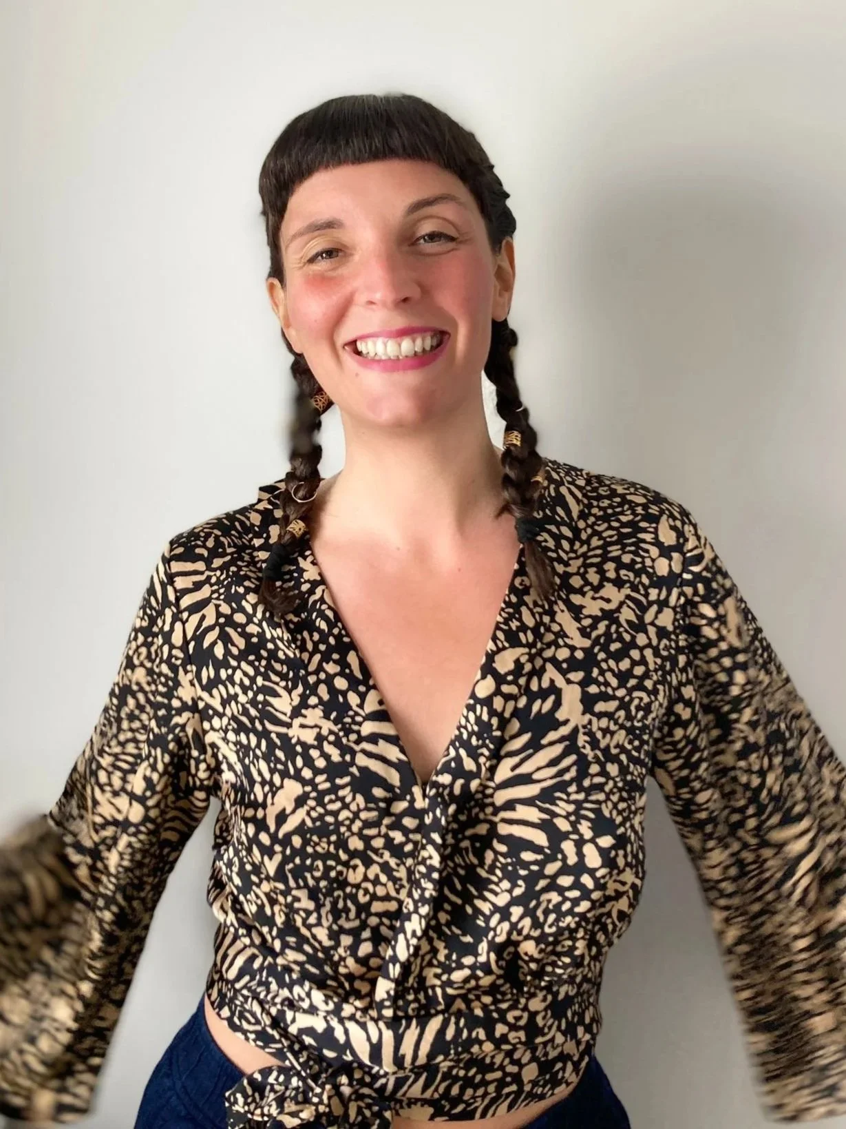 A smiling woman with braided brown hair, wearing a leopard print blouse, standing against a plain white wall.